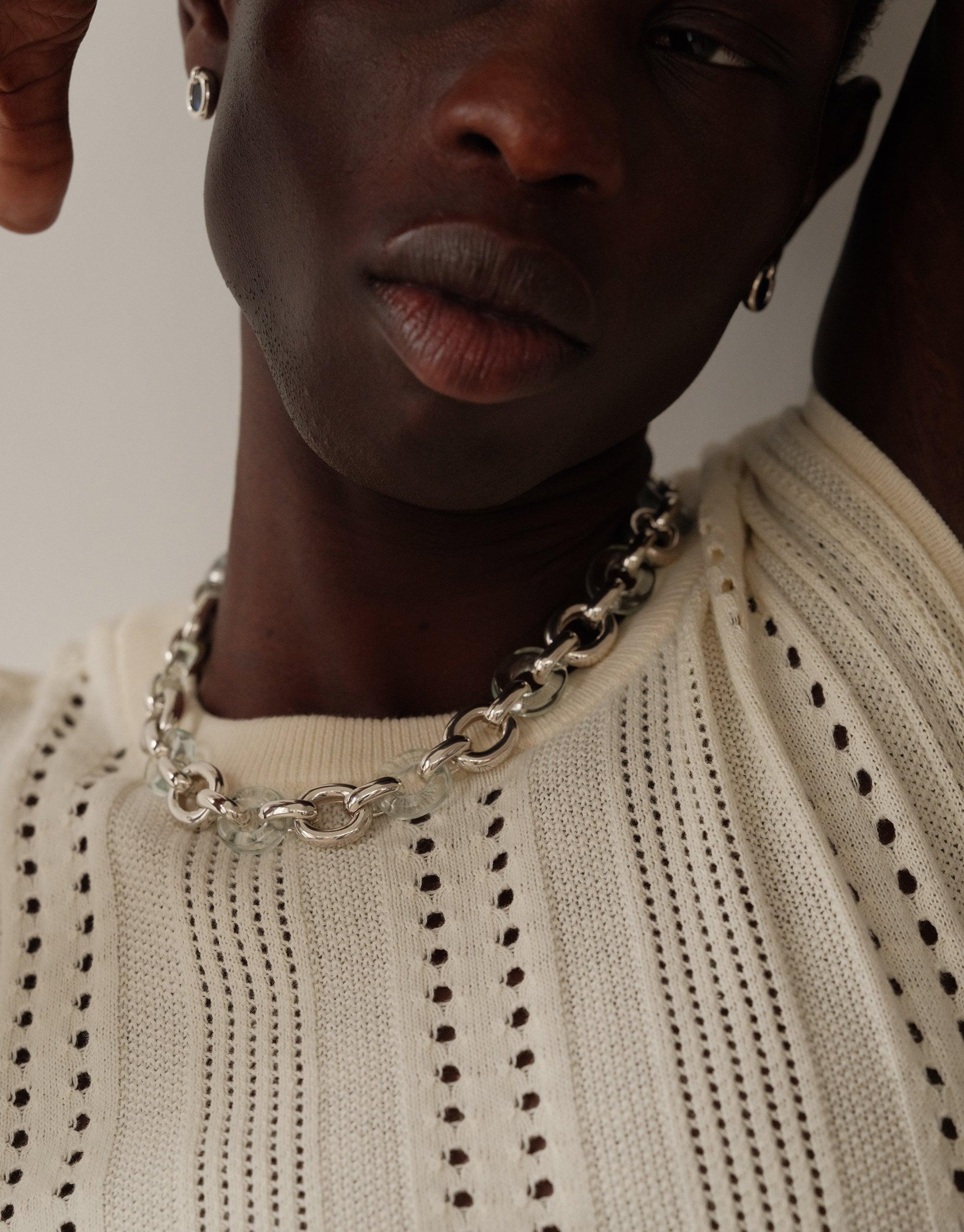 Close-up of a model wearing the Connected Loop Necklace in mixed metal, showcasing its intricate chain design against a cream-colored knit sweater. The necklace complements the textured garment, embodying unity and sustainable fashion. Connected Loop Necklace | Mixed Metal & Glass by CLED.