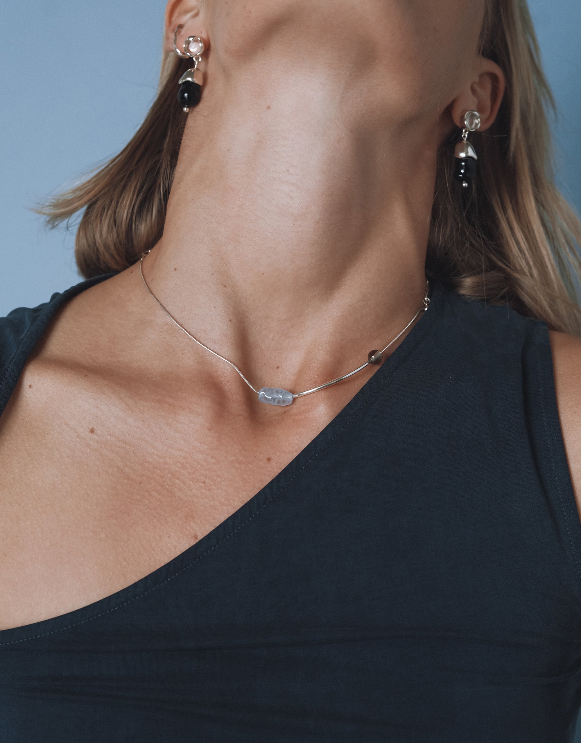 A woman wearing a dark, sleeveless, asymmetrical top is shown from the shoulders up against a blue background. She tilts her head back, revealing a silver choker necklace with one light blue glass bead in the center and two darker beads at each end. She also wears matching dangle earrings with round silver tops and black upcycled glass drops.
