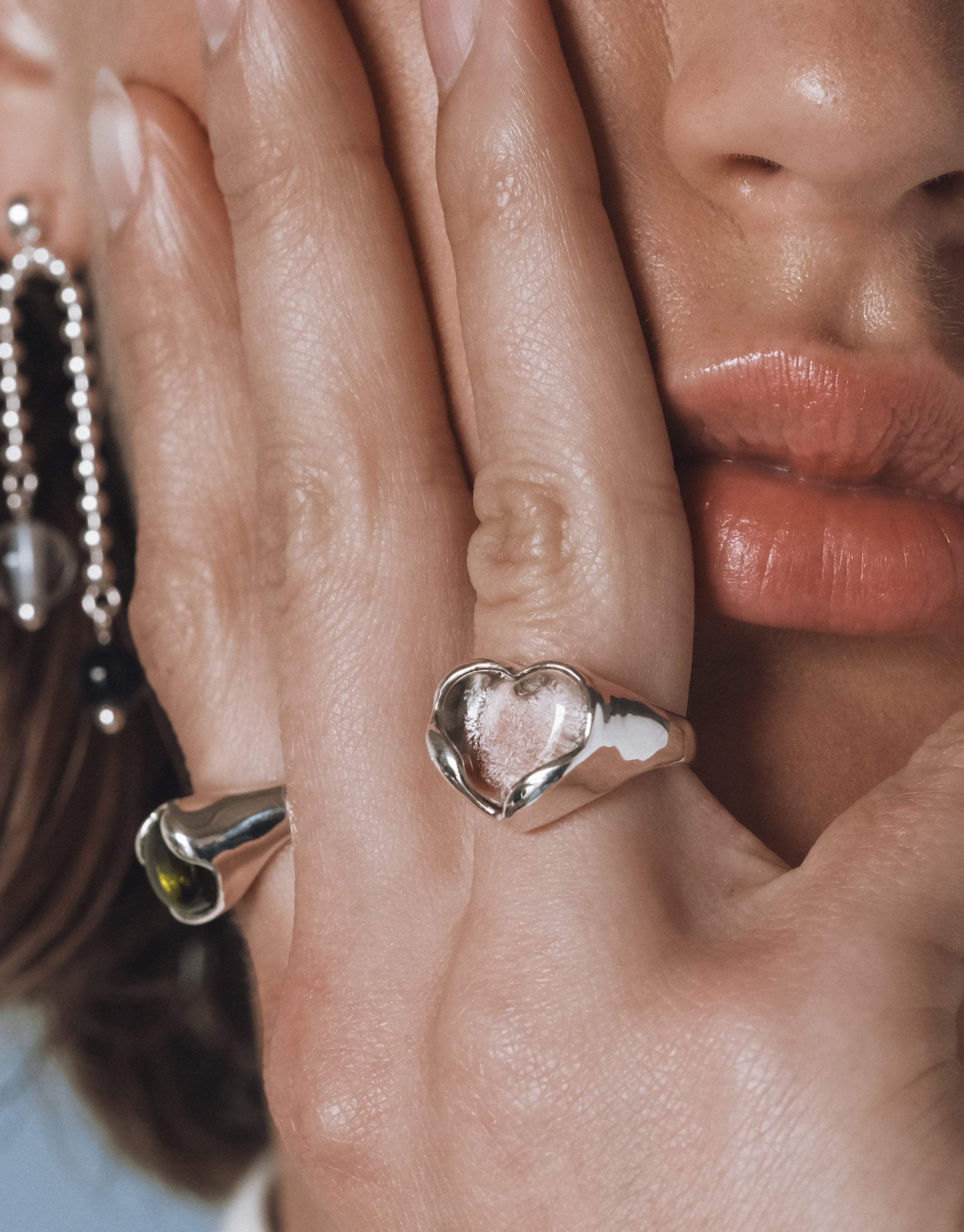 Close up of a model’s hand gently touching her face, showcasing a silver heart shaped ring with a clear Eco Gem and a sculptural silver ring with an olive green stone. A beaded earring with black and clear accents is visible in the background.