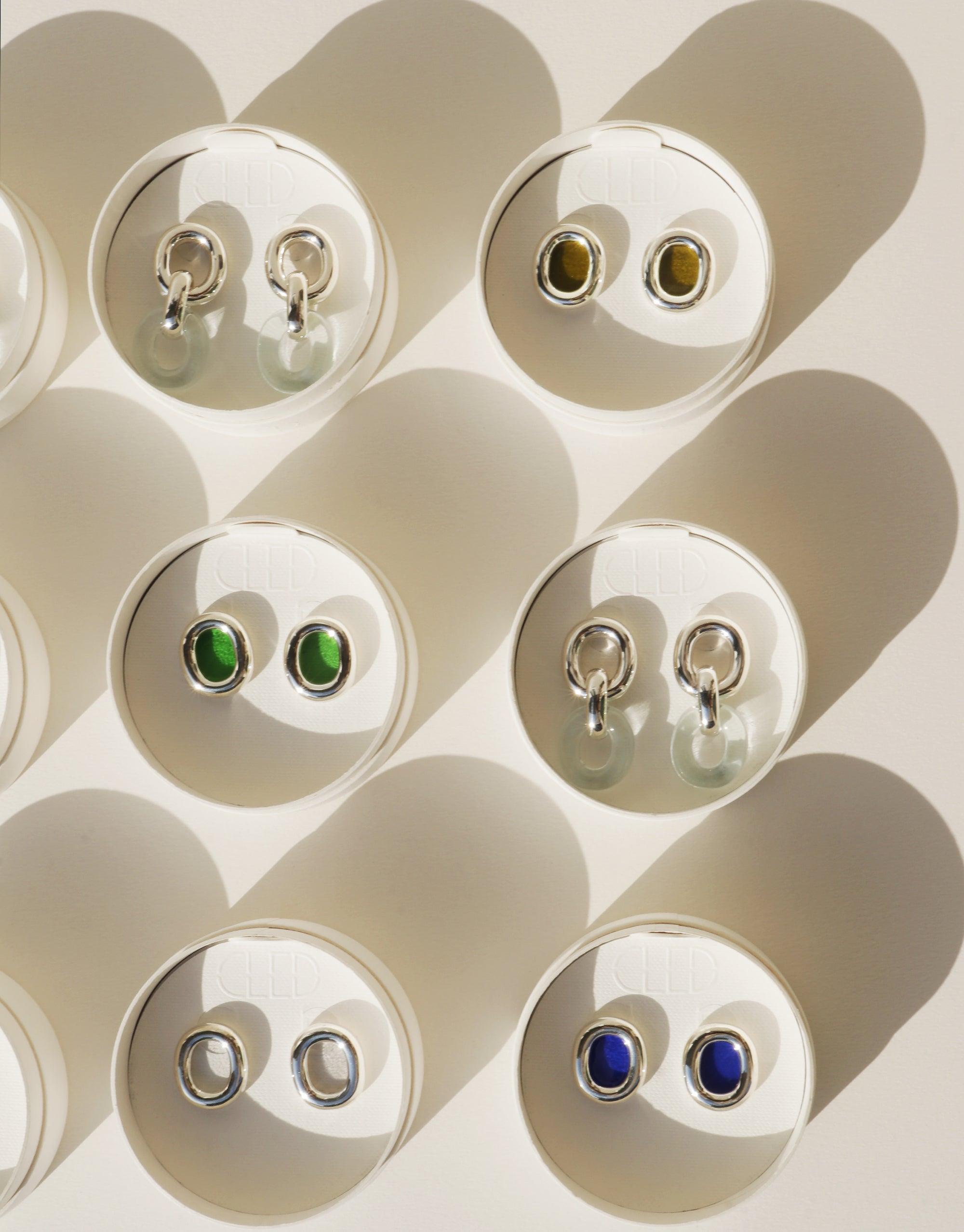 Assortment of Connected Loop Earrings displayed in circular cutouts on white background, featuring various colors and styles. Sustainable jewelry showcasing unity and upcycling. CLED Connected Loop Earrings.