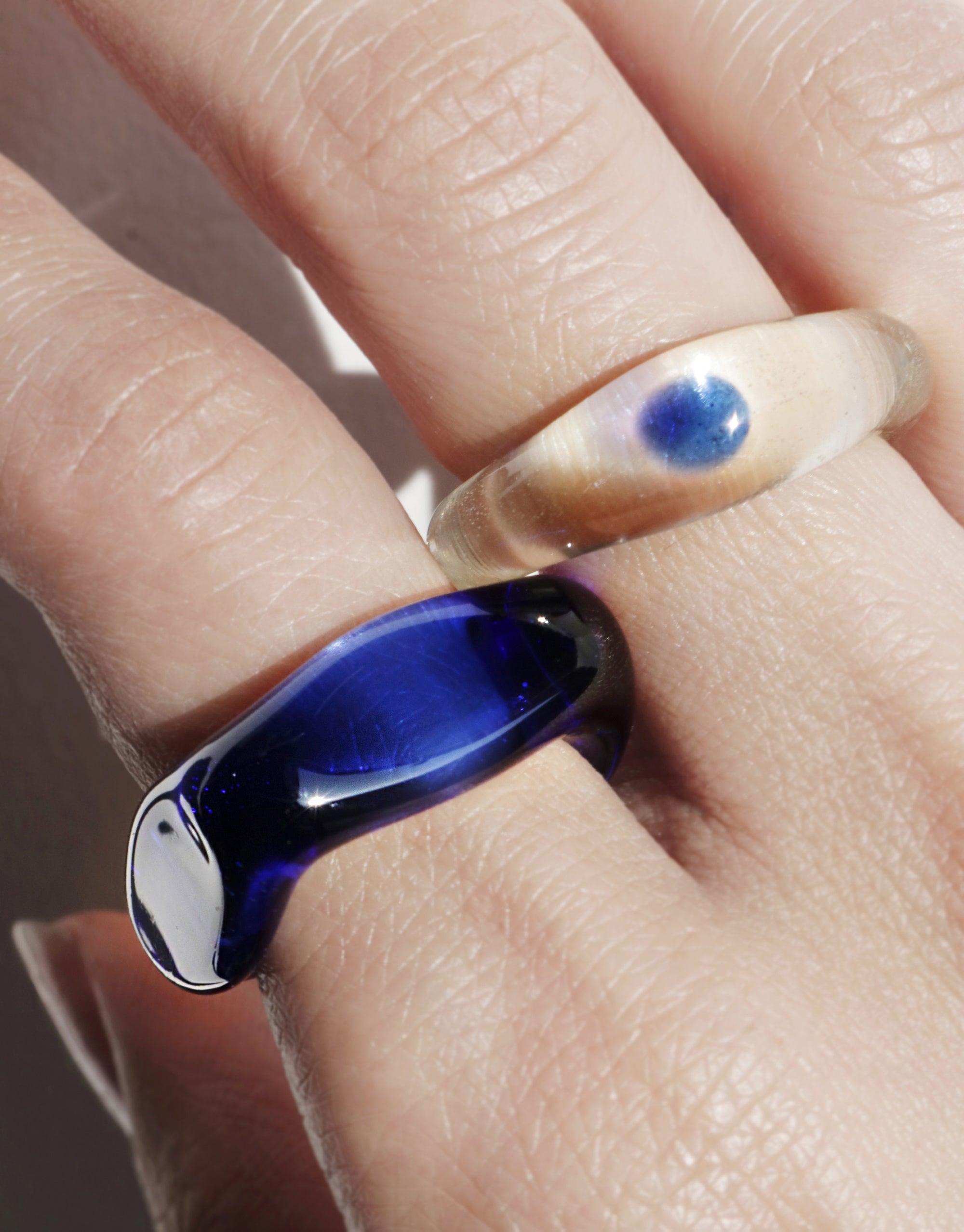 Close-up of two unique rings on a hand: a clear resin ring with a blue circular accent, and a sleek cobalt blue ring with a curved design. The Blue Moon Ring showcases elegant craftsmanship and celestial-inspired beauty. CLED