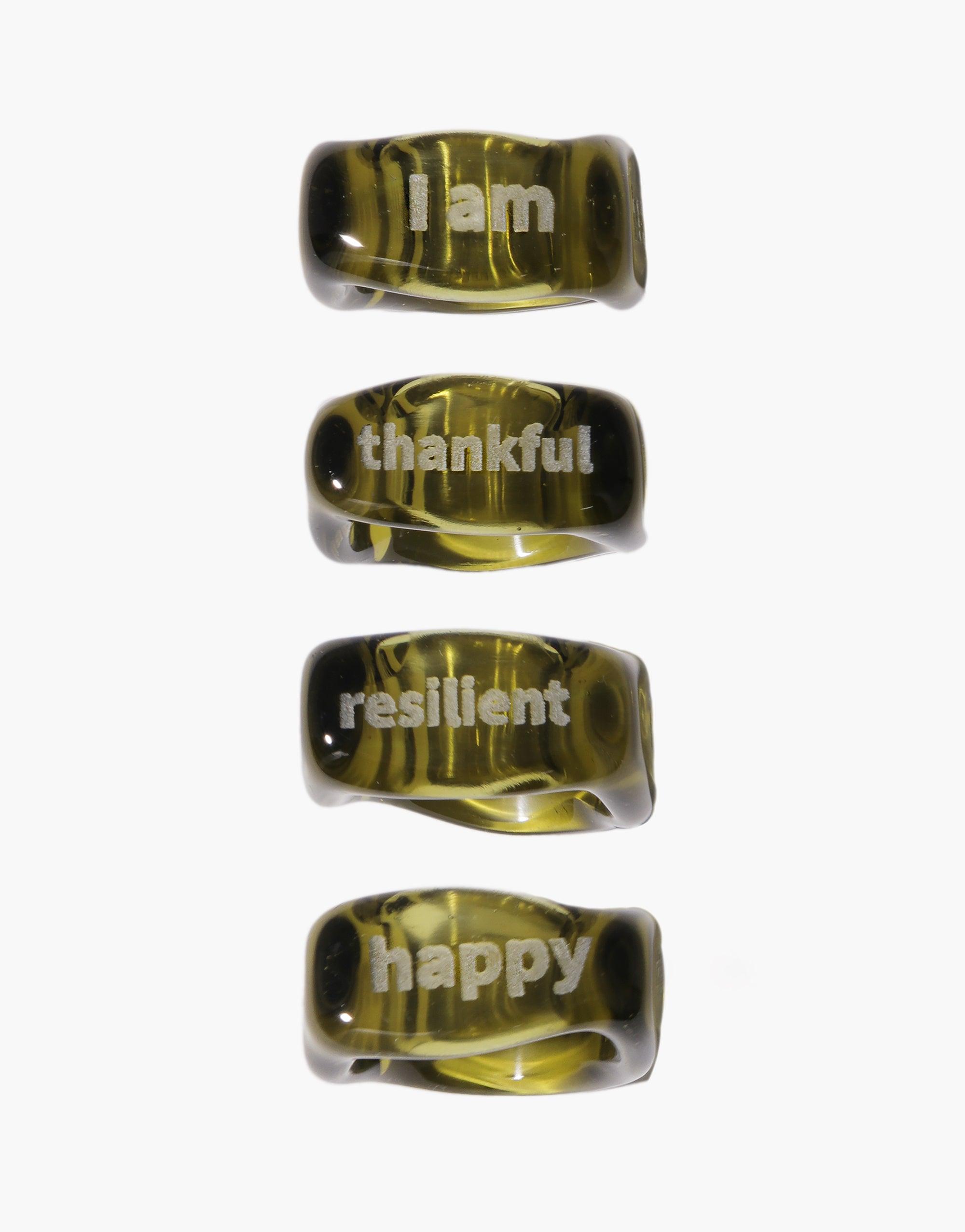 Four square-shaped glass rings with engraved positive affirmations: "I am", "thankful", "resilient", and "happy". Unique molten design with a yellow-green tint. Personalized jewelry for self-empowerment and everyday reminders. Dice Ring | I am by CLED.
