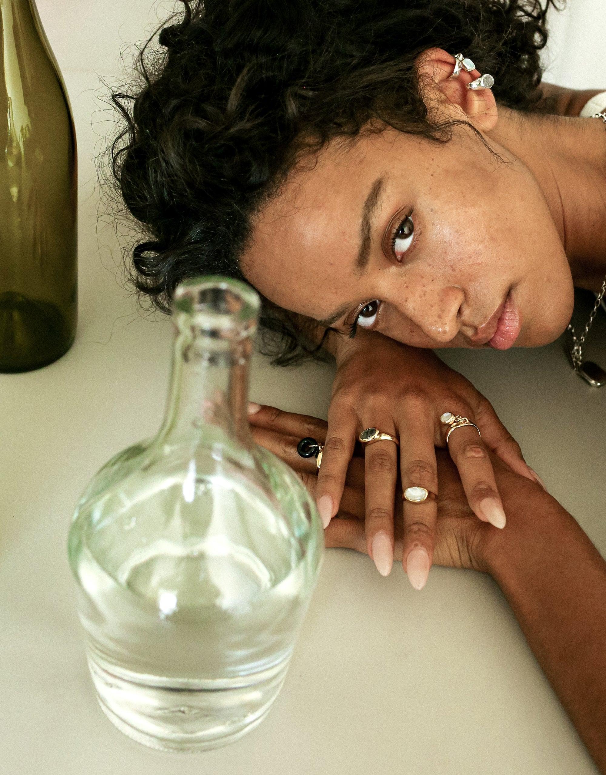 Model showcasing Oval Sculpture Ring, lying on white surface with empty glass bottles. Hands displaying multiple rings, emphasizing eco-friendly jewelry design. Artistic composition highlighting sustainable fashion and upcycled materials. Oval Sculpture Ring by CLED.