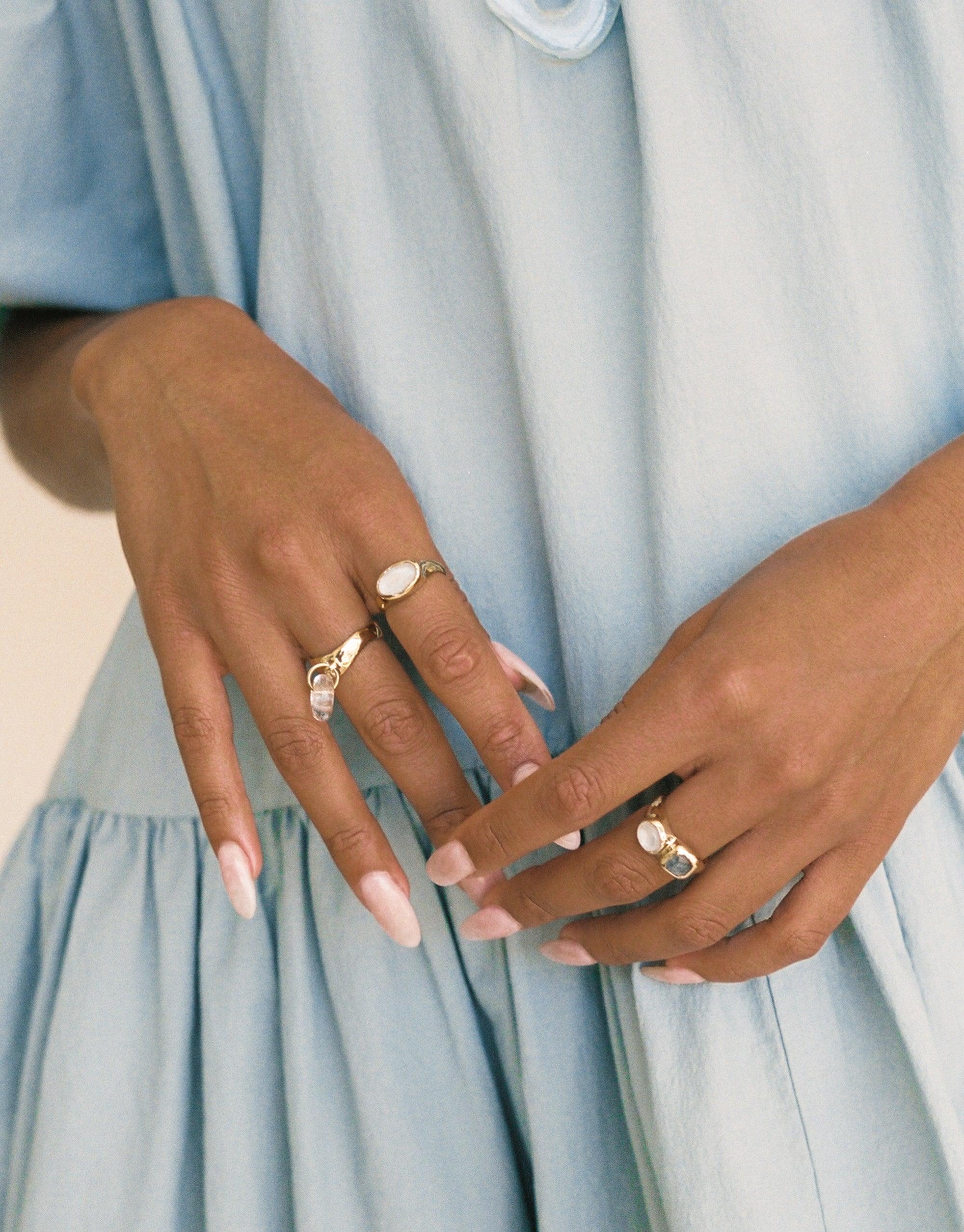 Hands wearing multiple delicate gold rings with oval white stones, showcasing the Oval Sculpture Ring. Elegant jewelry displayed on fingers against a light blue dress background. Handcrafted eco-friendly accessories by CLED.
