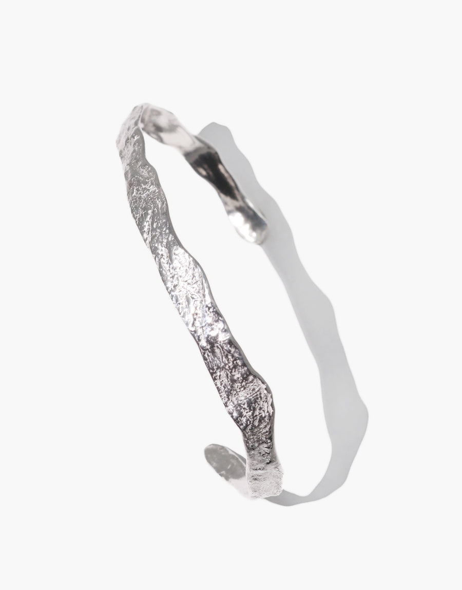 A silver bracelet with a raw, organic texture and irregular shape, resembling a rugged cliff or natural terrain. The bracelet casts a soft shadow on a light background, highlighting its reflective surface and sculptural form.