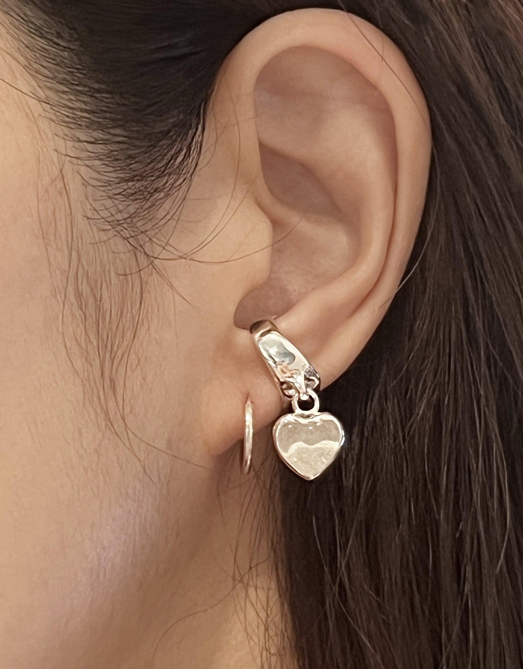 Close-up of an ear wearing a delicate Heart Dangling Ear Cuff in silver, featuring a small heart-shaped charm hanging from a thin cuff. The cuff wraps around the upper part of the ear, creating a unique and stylish look. Handcrafted jewelry by CLED.