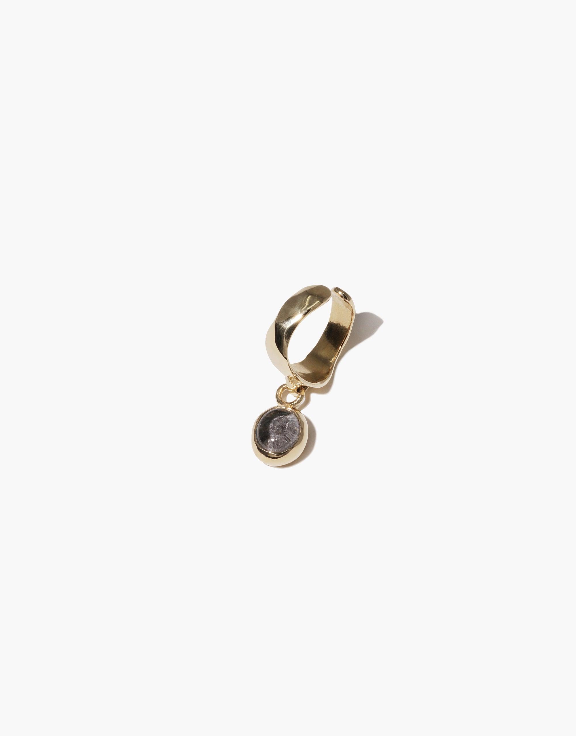 Oval Dangling Ear Cuff featuring a gold-toned metal cuff with a small circular charm pendant. The pendant contains a dark-colored upcycled glass element, showcasing sustainable jewelry design. Handcrafted elegance for eco-conscious fashion lovers. CLED, Oval Dangling Ear Cuff.