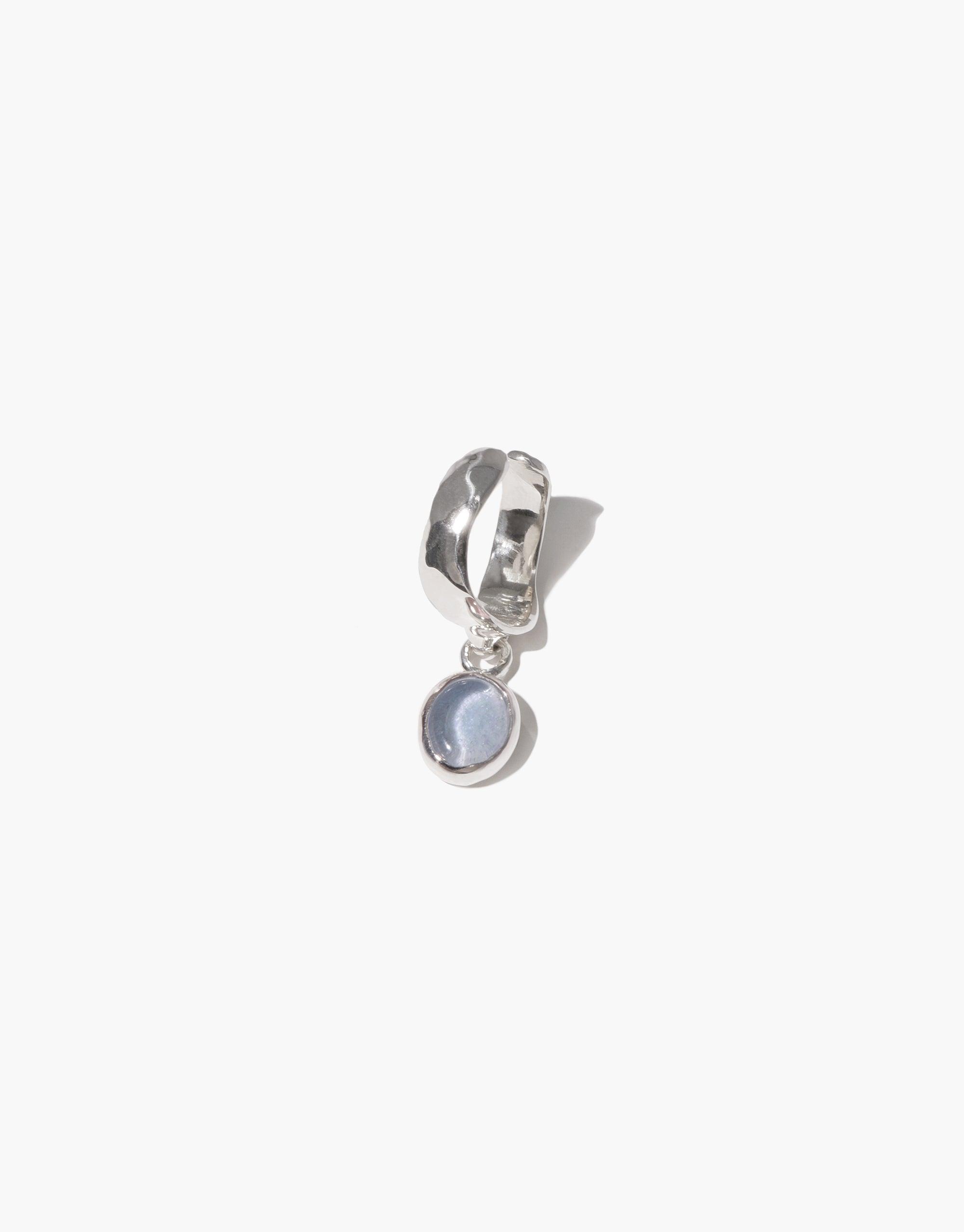 Oval Dangling Ear Cuff featuring a silver cuff with a small, round, light blue charm hanging from it. Handcrafted jewelry piece made from recycled materials, showcasing delicate craftsmanship and eco-friendly design. CLED