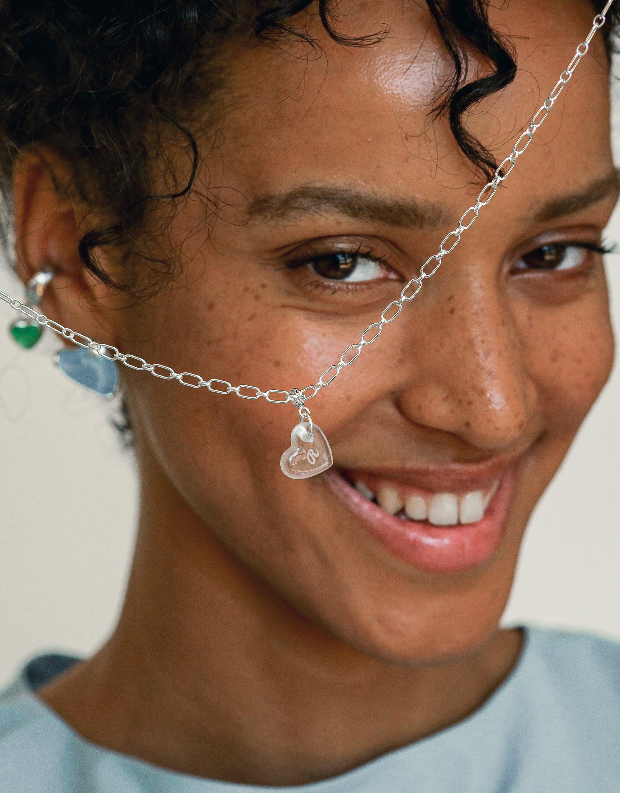 Close-up of silver chain necklace with delicate heart-shaped charm, showcasing the Heart Icon Charm. Necklace rests elegantly on model's skin, highlighting the charm's intricate design and versatile appeal. Sustainable jewelry perfect for layering or wearing solo. CLED, Heart | Icon Charm.
