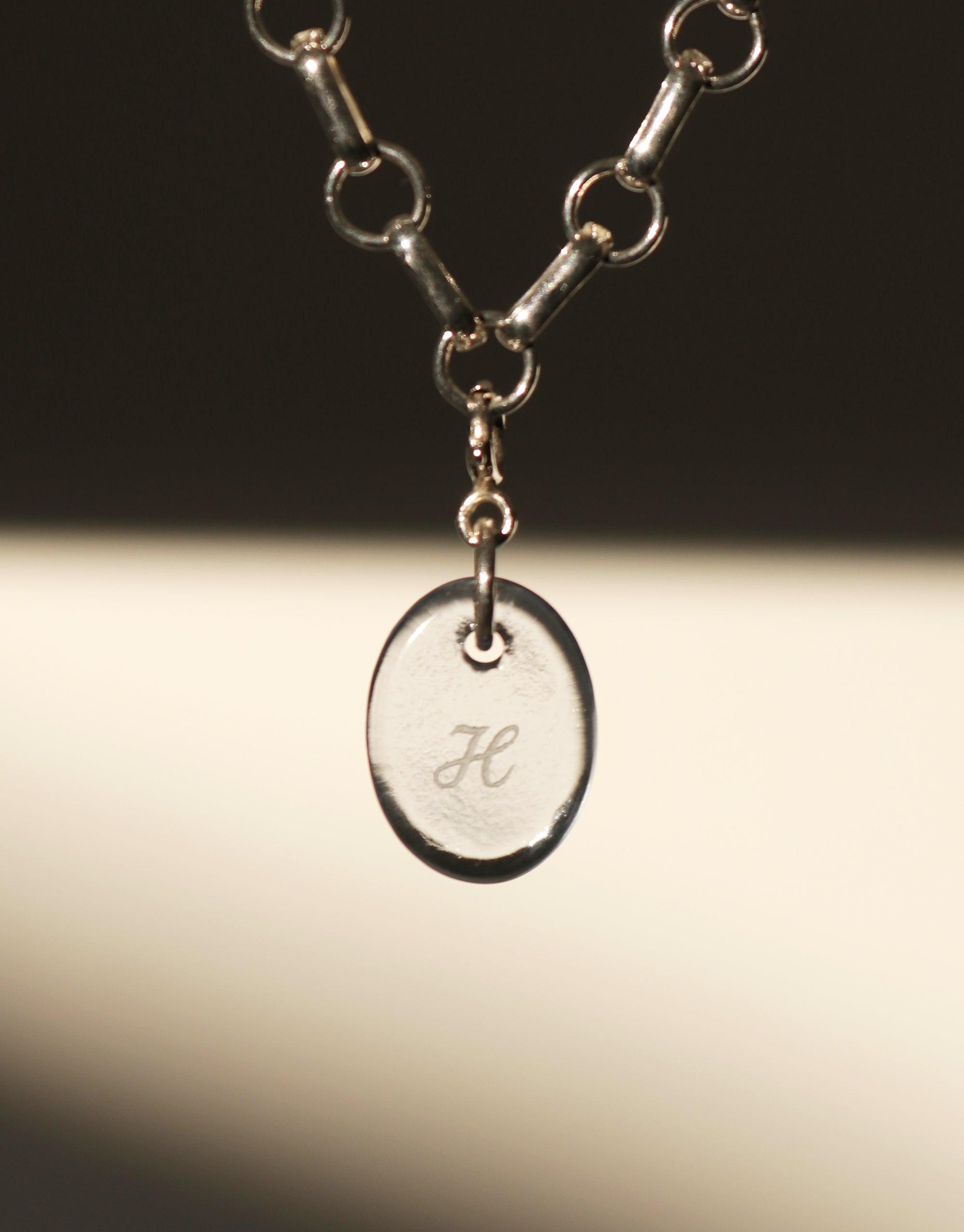 Close-up of a delicate silver chain necklace featuring an oval-shaped pendant charm with an engraved letter 'H'. The pendant hangs from a sturdy link chain, showcasing the Oval Icon Charm's elegant simplicity and customizable design. Handcrafted jewelry by CLED.