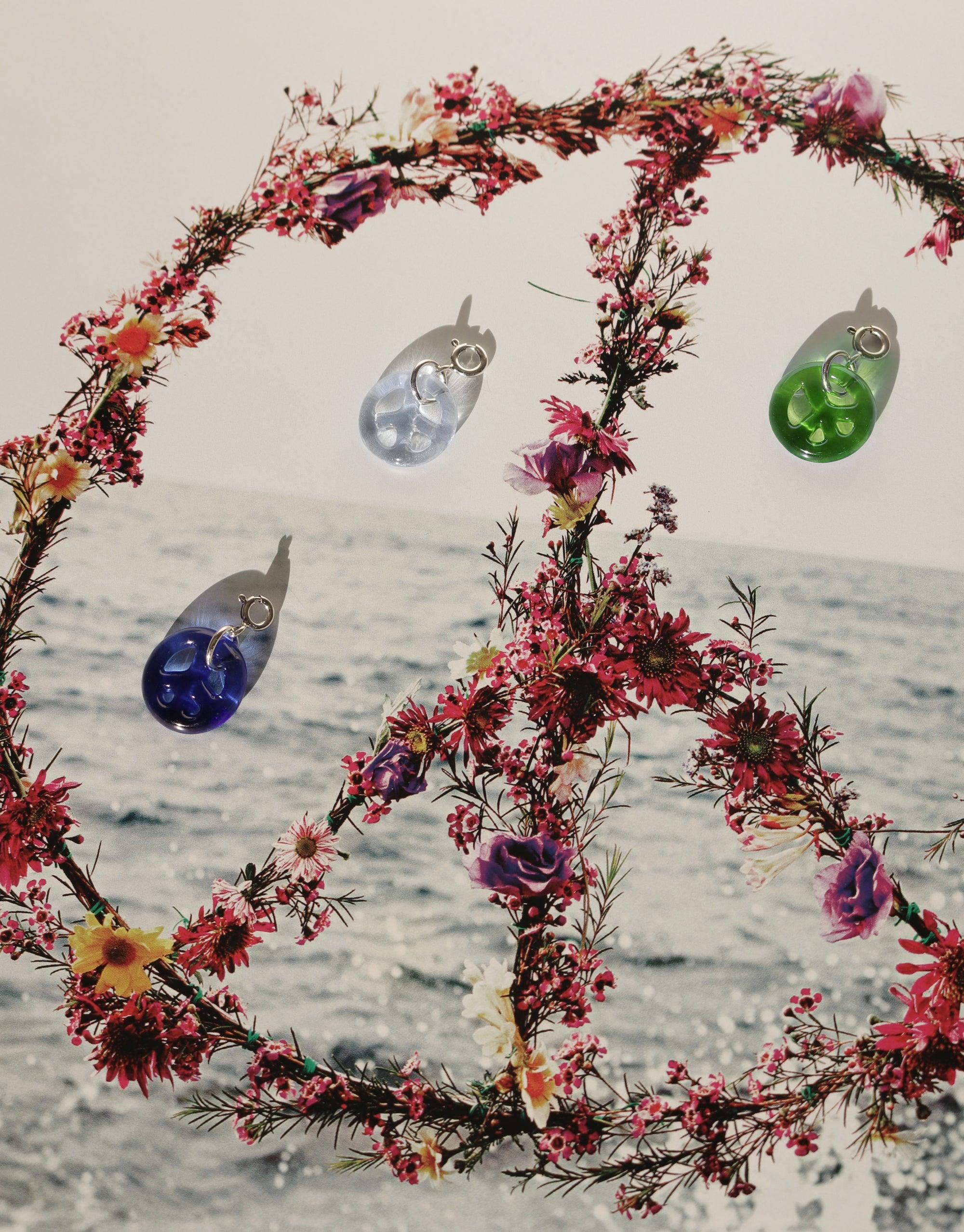 Peace Icon Charms in various colors hanging from floral peace sign wreath against ocean backdrop, showcasing eco-friendly glass jewelry. Handcrafted sustainable accessories for necklaces and bracelets. Peace | Icon Charm by CLED