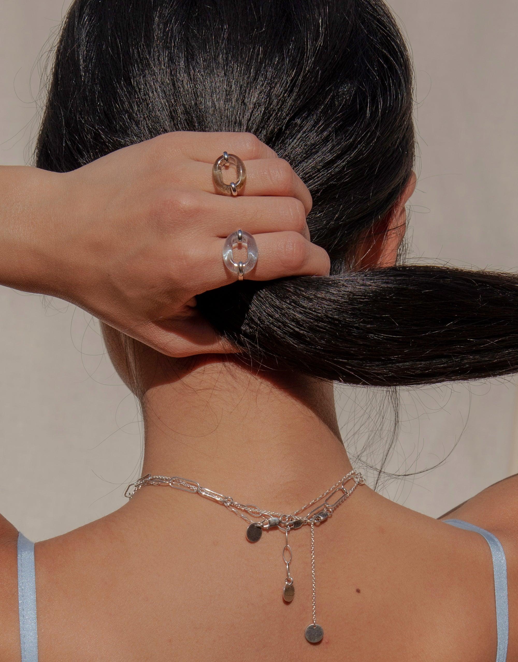 Close-up of a hand wearing two In The Loop Rings, showcasing their unique circular design. The rings are displayed on fingers holding dark hair, with a delicate necklace visible on the neck. Sustainable and stylish jewelry by CLED.