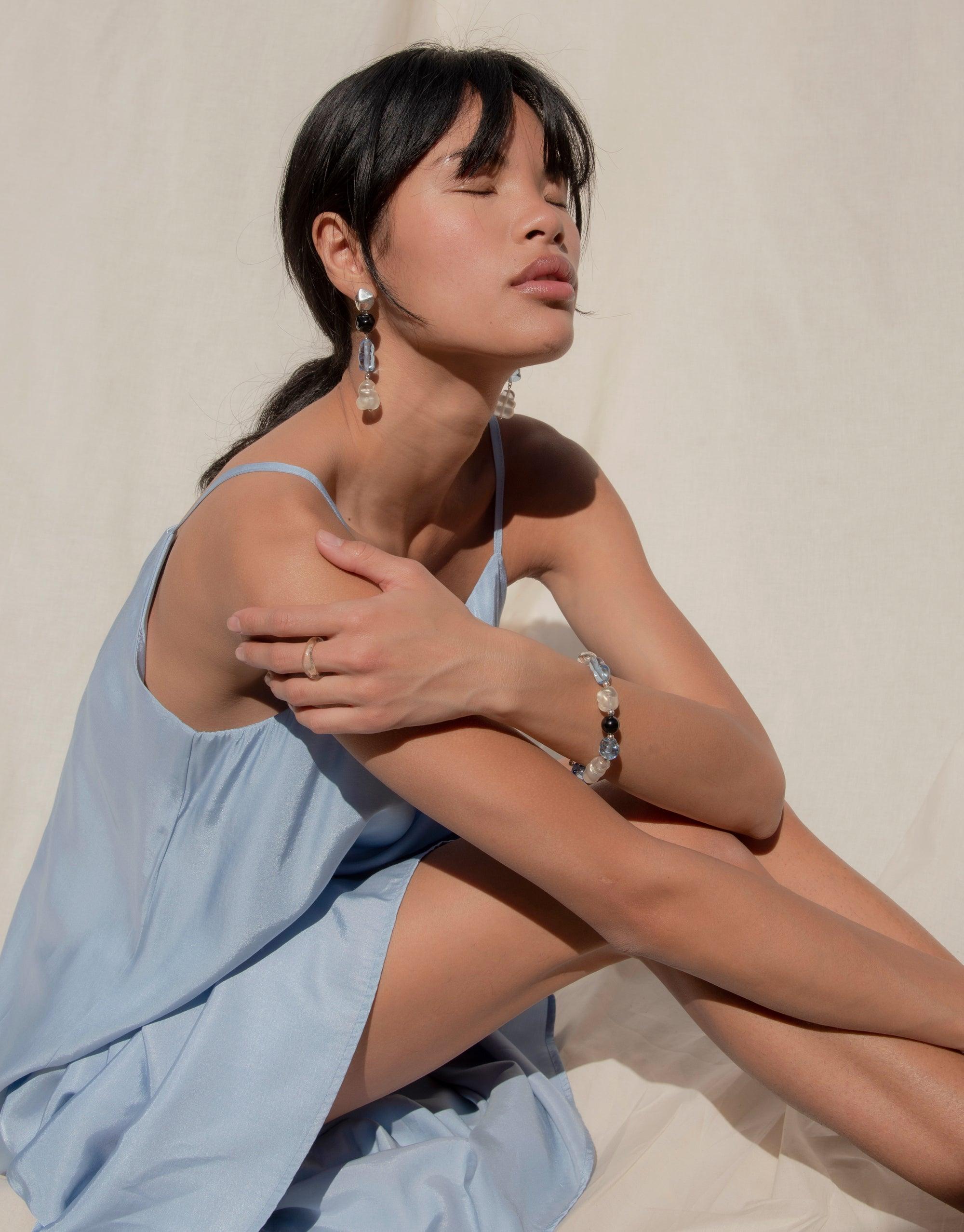 Woman wearing light blue dress showcasing elegant Blue Pearl Earrings with multi-color swaying drops, inspired by Blue Jay bird. Handcrafted eco-friendly jewelry made from repurposed glass bottles. Sustainable fashion accessory. Blue Pearl Earrings by CLED.