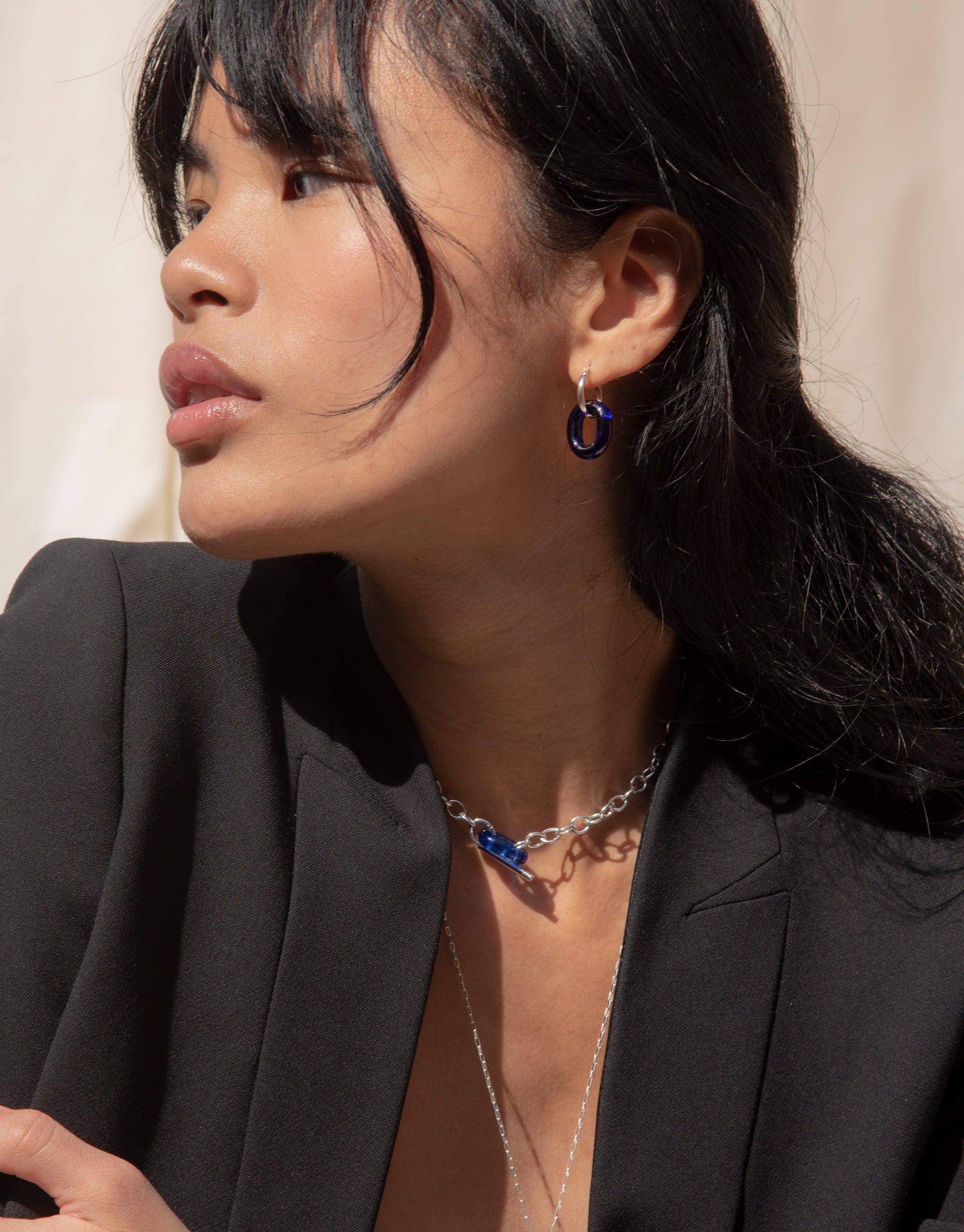Close-up of a woman wearing small hoop earrings with blue eco gems, showcasing the "In The Loop Hoops | Small" design. The earrings complement her elegant profile and black blazer, highlighting the versatility and style of CLED's sustainable jewelry.