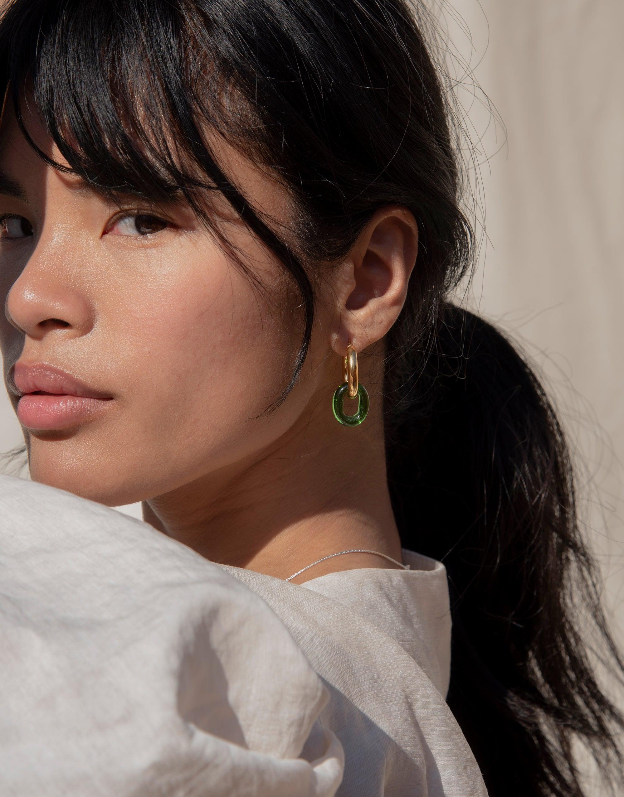 Close-up of a woman wearing large gold hoop earrings with a small green eco gem loop. The earrings showcase the "In The Loop Hoops | Large" design, highlighting the lightweight hollow tubing and signature eco gem made from recycled glass. Handcrafted sustainable jewelry by CLED.