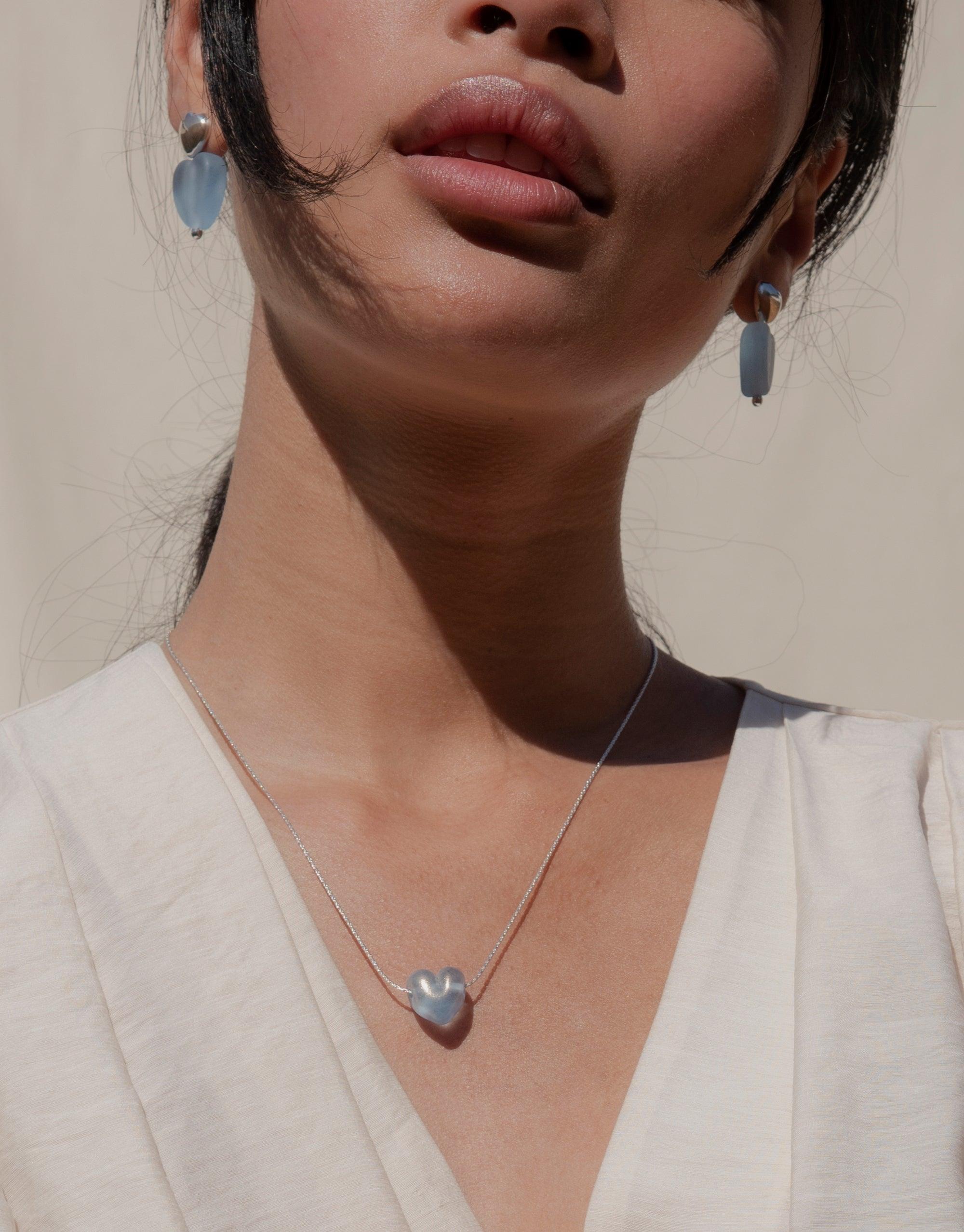 Elegant matte blue heart-shaped earrings and matching necklace on a model, showcasing the Love Earrings with Large Heart Drop. Handcrafted eco-friendly jewelry made from repurposed glass, highlighting sustainable fashion by CLED.