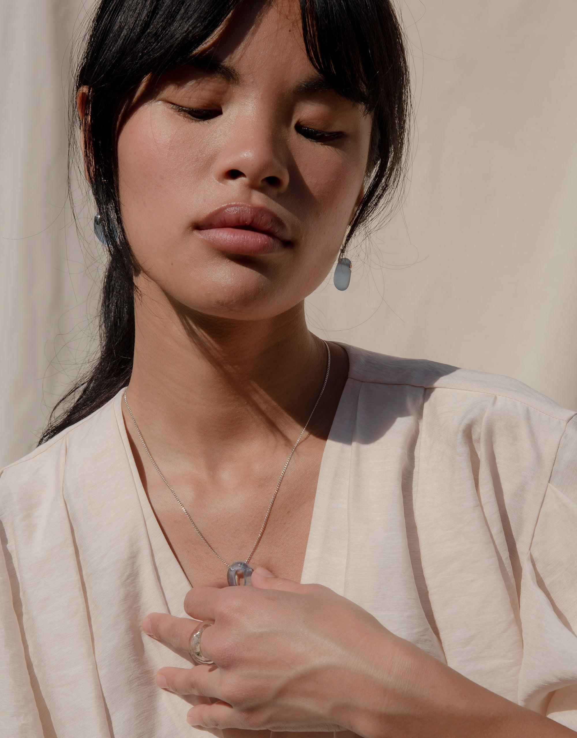 Elegant model showcasing Pebble Earrings, handcrafted eco-friendly jewelry featuring matte-finished glass drops. Sustainable luxury accessory paired with delicate necklace and ring. Soft lighting highlights the earrings' unique shape and luminous quality. Ethically made in Los Angeles by CLED.