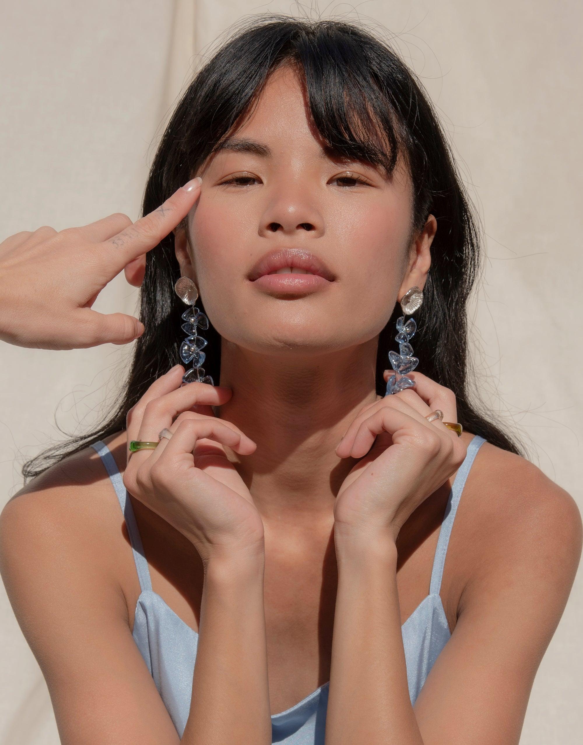 Close-up of a model wearing long, delicate earrings with light blue glass petal drops. The earrings showcase an elegant, nature-inspired design that complements the model's soft blue top. Periwinkle Trio Earrings by CLED.