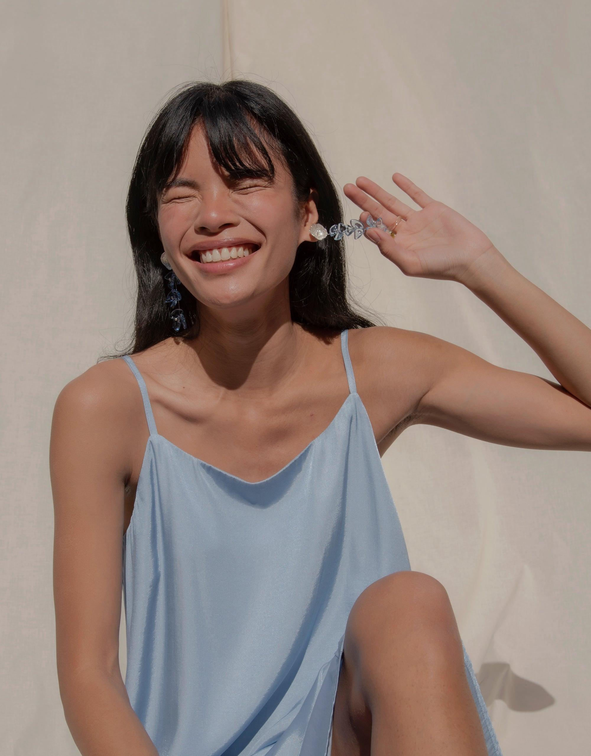 Woman in light blue dress smiling and touching earring, showcasing Periwinkle Trio Earrings with delicate flower studs and glass petal drops. Handcrafted eco-friendly jewelry by CLED