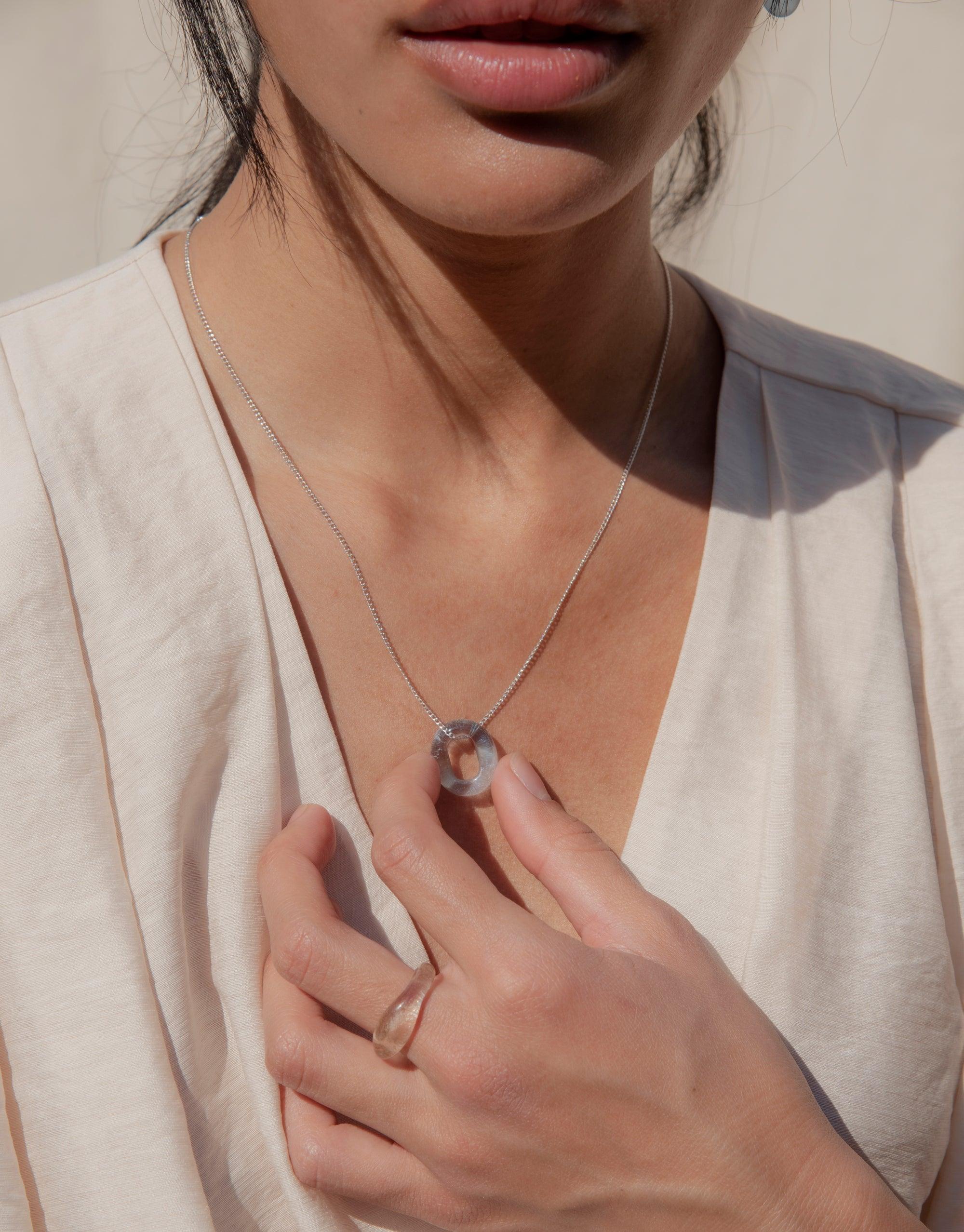 Close-up of a person wearing a delicate silver chain necklace with a small, circular eco glass pendant. The necklace rests against a light-colored top, showcasing the minimalist design of the In The Loop Necklace by CLED.