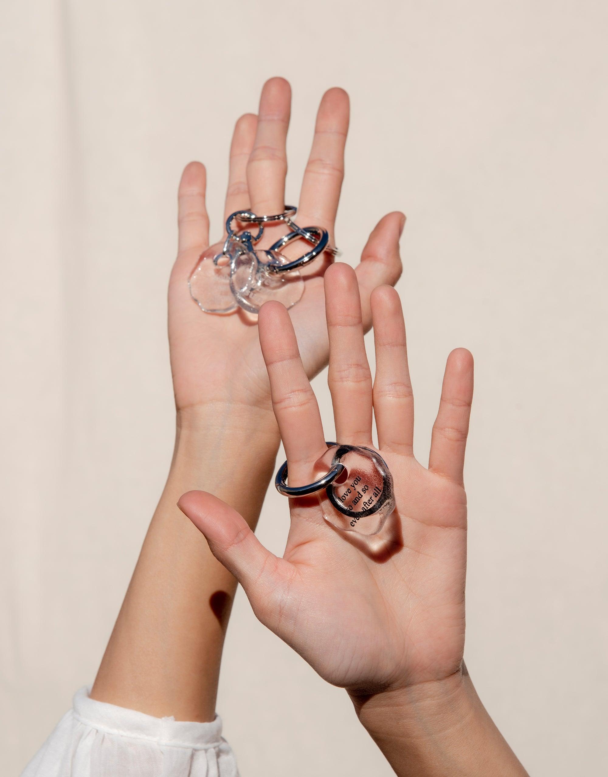 Hands holding Messenger Keyring | Love with clear glass charm and metal clasp. Eco-friendly keyring featuring engraved love message on repurposed glass. Handmade accessory for expressing affection and connection. Sustainable jewelry by CLED.