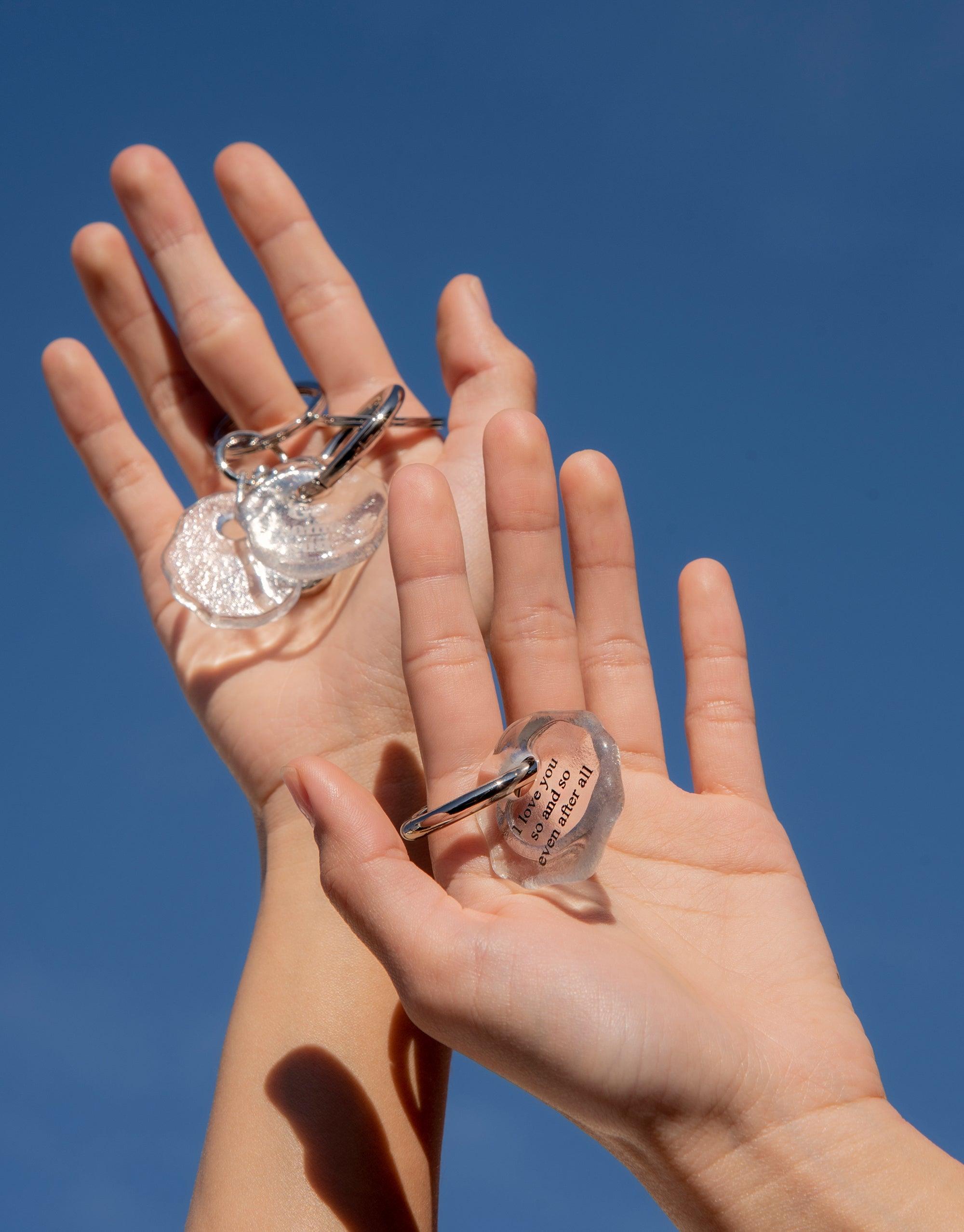 Hands holding eco-friendly glass keyrings with inspirational messages against blue sky background. Messenger Keyring with "Look at the sky" engraving showcases sustainable, handmade design. Perfect for sharing thoughts and connecting with loved ones. CLED. Messenger Keyring | Look at the sky