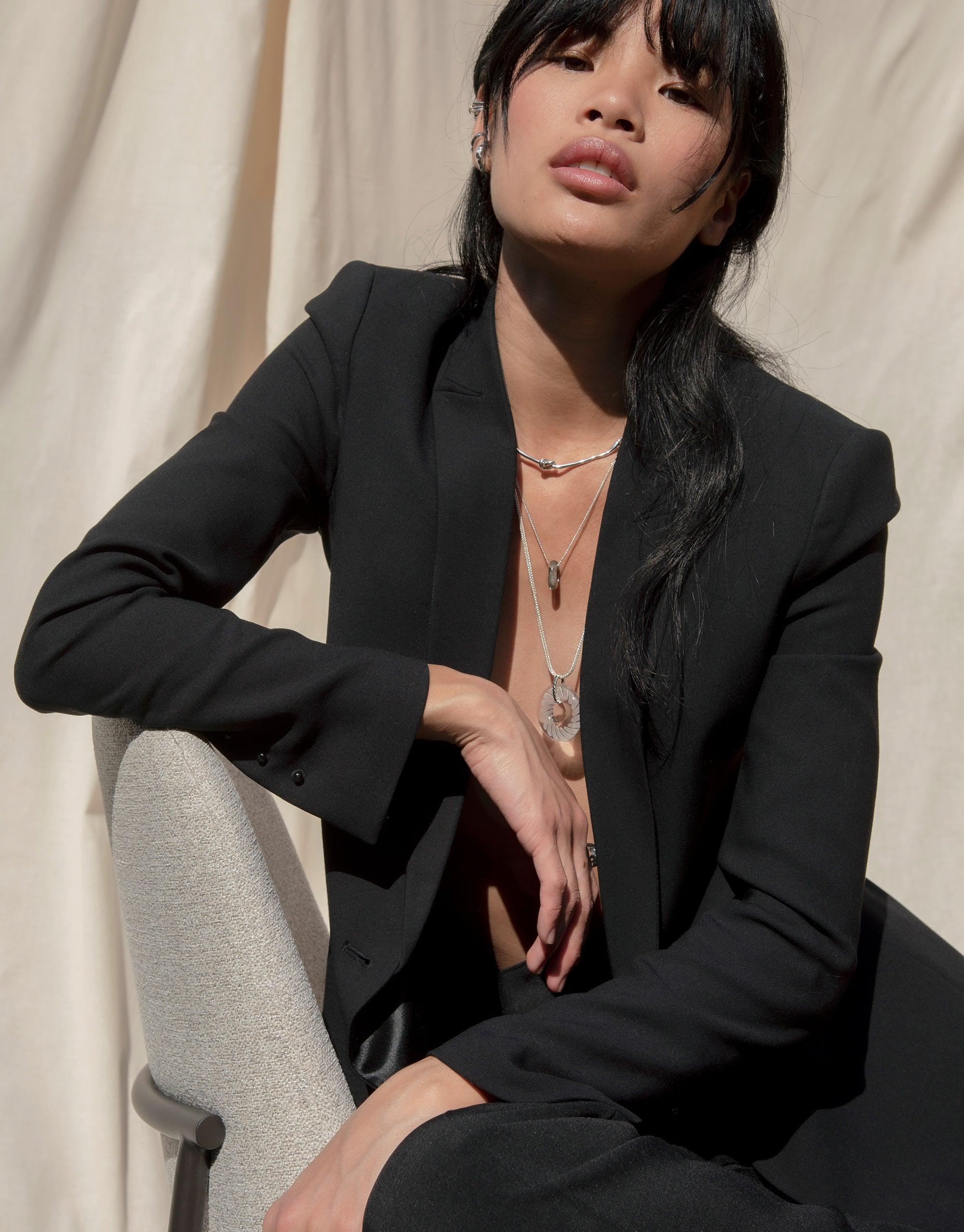 Woman in black blazer wearing delicate layered necklaces, showcasing elegant jewelry design against neutral background. Braid Necklace by CLED visible among the layers, highlighting the product's versatile and stylish appeal.