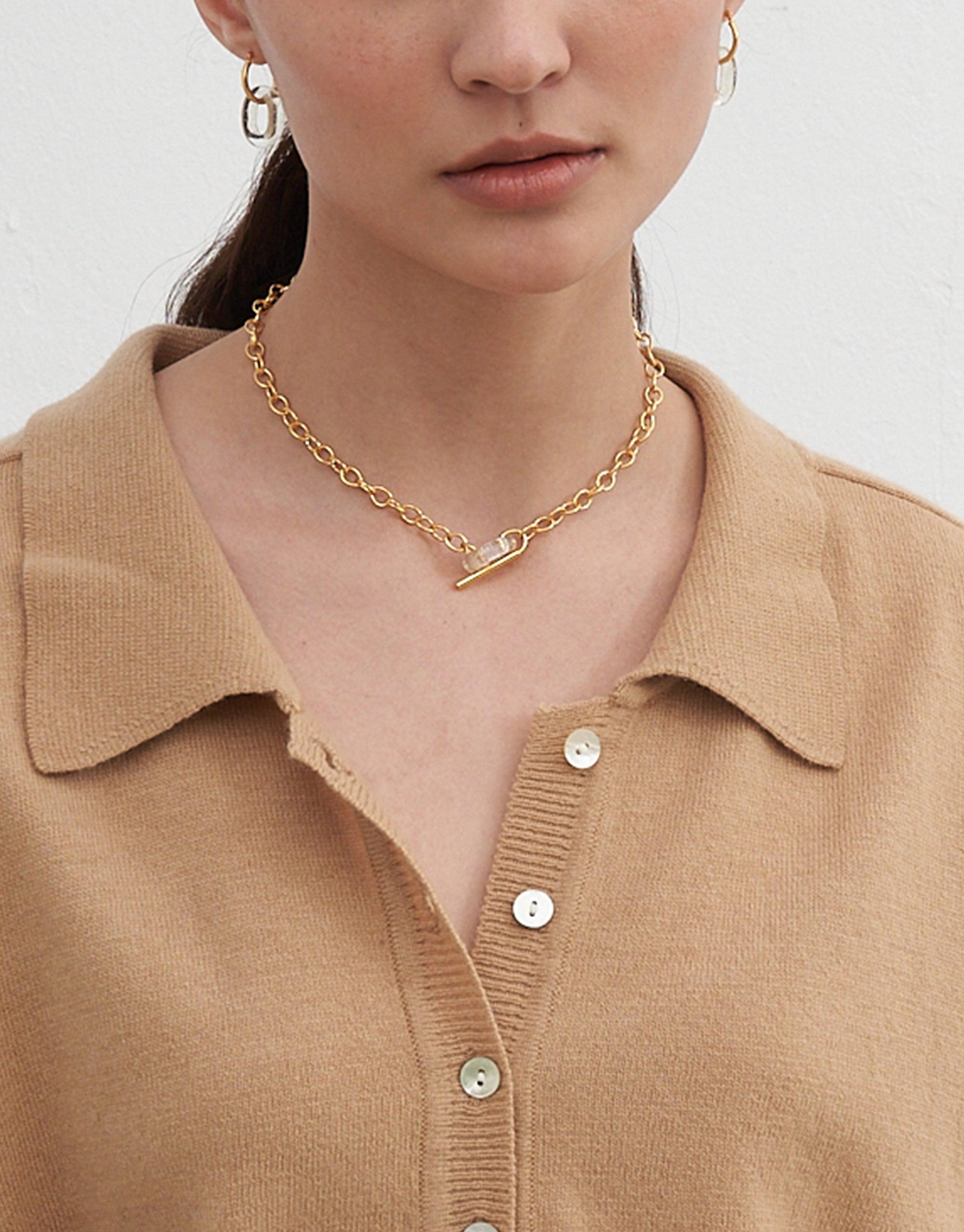 Close-up of a person wearing a gold ITL Toggle Choker Necklace with interlocking links and a small loop eco gem. The necklace sits elegantly on the neck, complementing a beige collared sweater. Showcases the versatile and stylish design of the sustainable jewelry piece by CLED.