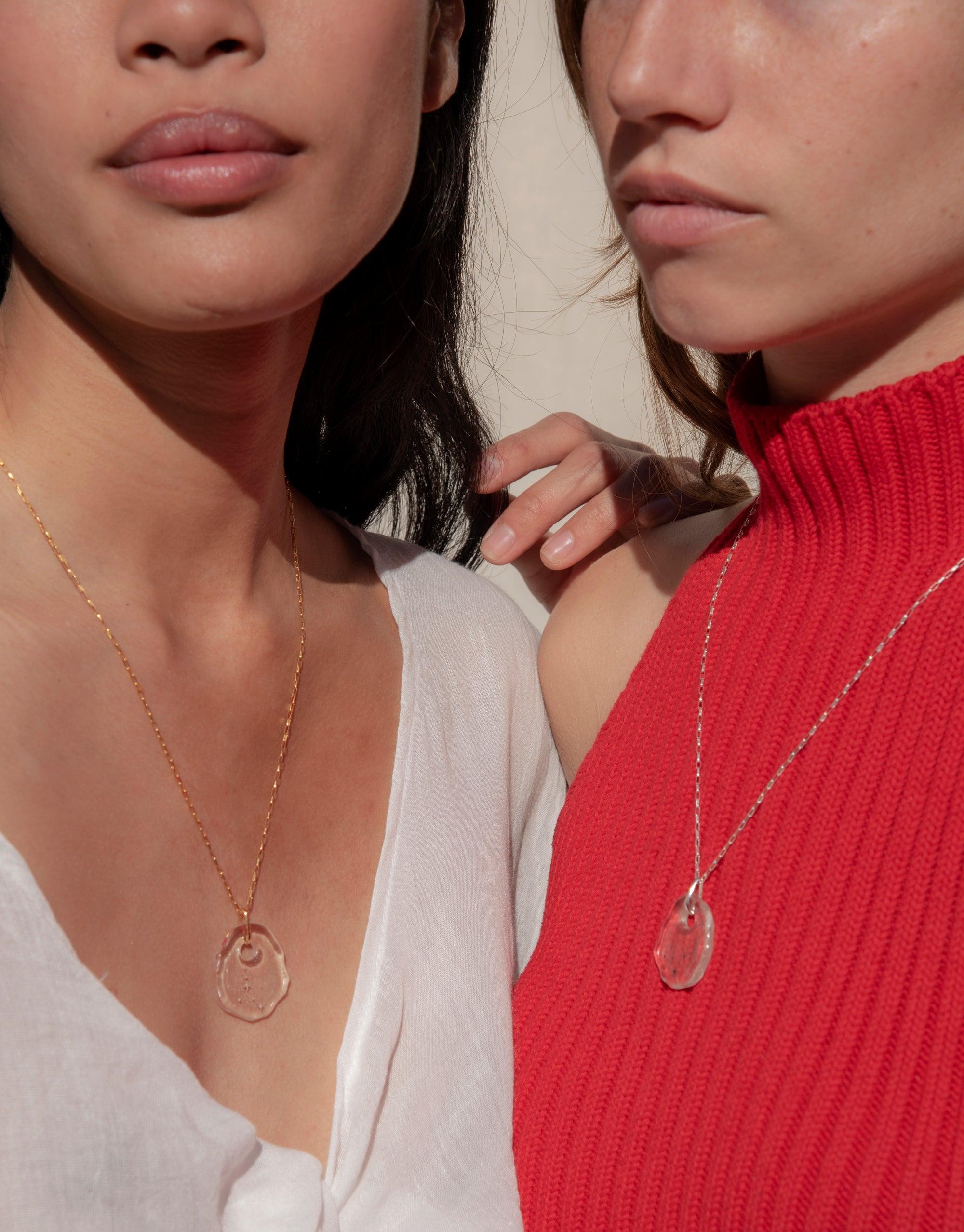 Two models wearing Constellation Necklaces with clear glass pendants on gold and silver chains. One in white top, one in red sweater. Showcasing versatile styling of eco-friendly zodiac-inspired jewelry. Handcrafted Messenger Collection by CLED.