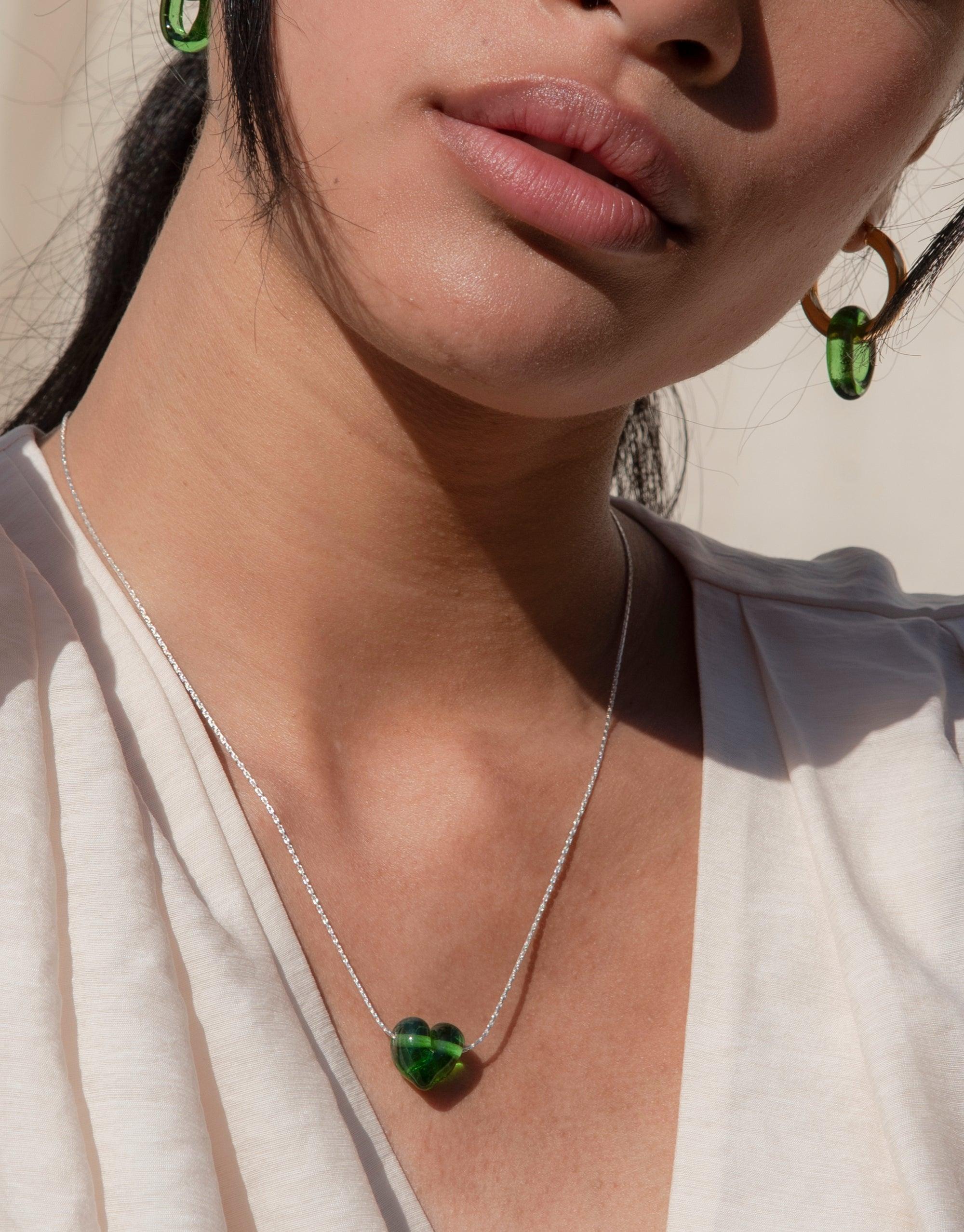 Close-up of a person wearing In The Loop Hoops | Large, featuring green eco gems. The image showcases the hoops' unique design and the complementary green heart-shaped pendant necklace, highlighting CLED's sustainable jewelry collection.