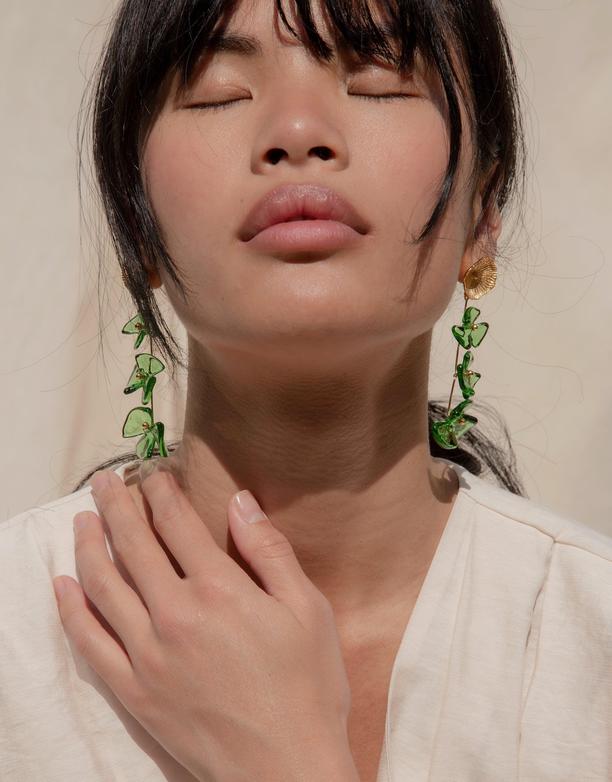 Close-up of a person wearing elegant green drop earrings with delicate leaf-like shapes, showcasing the Periwinkle Trio Earrings. The earrings hang gracefully, highlighting their unique eco-friendly design and handcrafted quality. Perfect for nature-inspired, sustainable jewelry enthusiasts. CLED