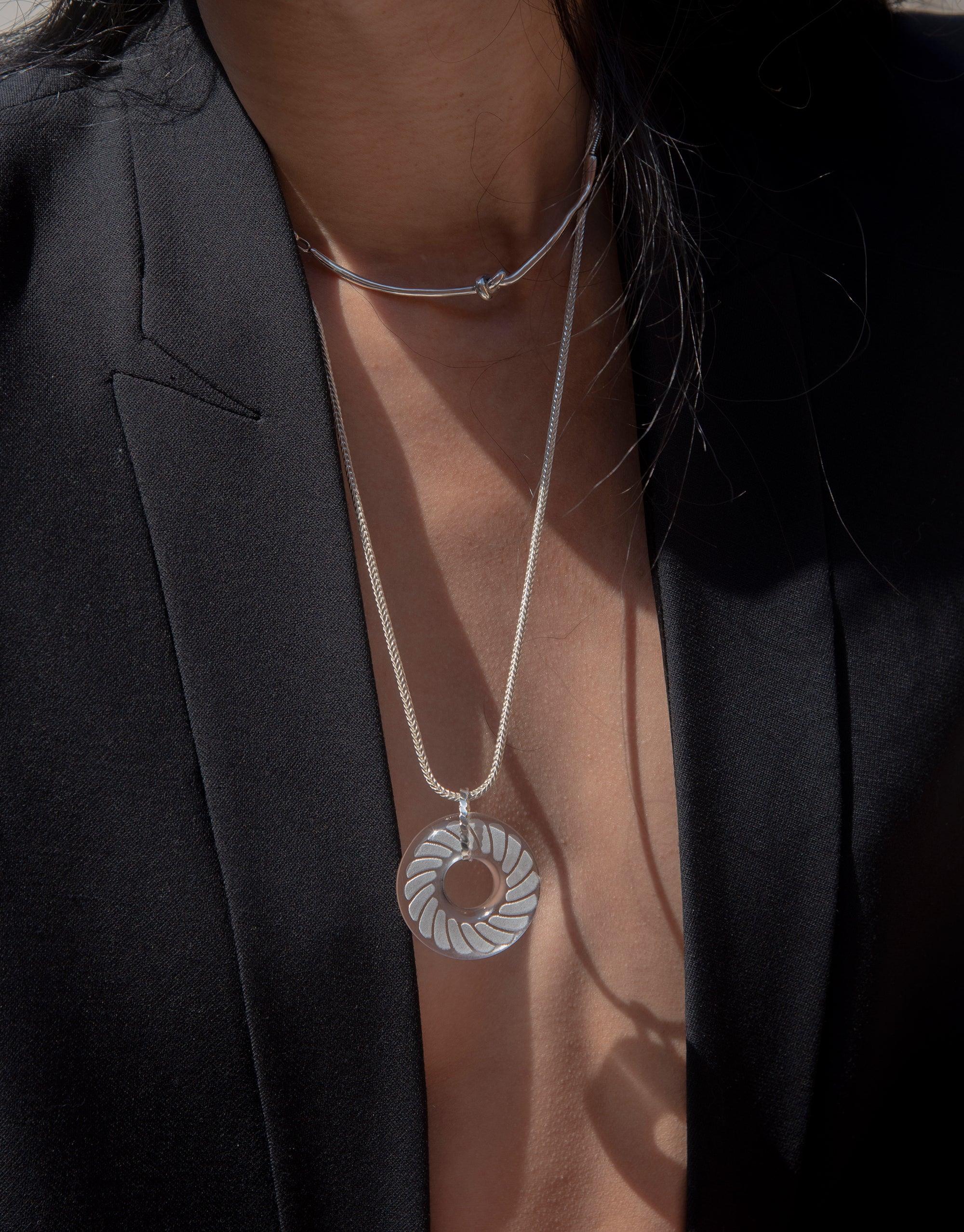 Elegant Braid Necklace with circular eco gem pendant on silver chain, worn with black blazer. Handcrafted torus-shaped pendant features intricate braid pattern engraving. Sustainable jewelry piece showcasing unique design and eco-friendly materials. CLED Braid Necklace.