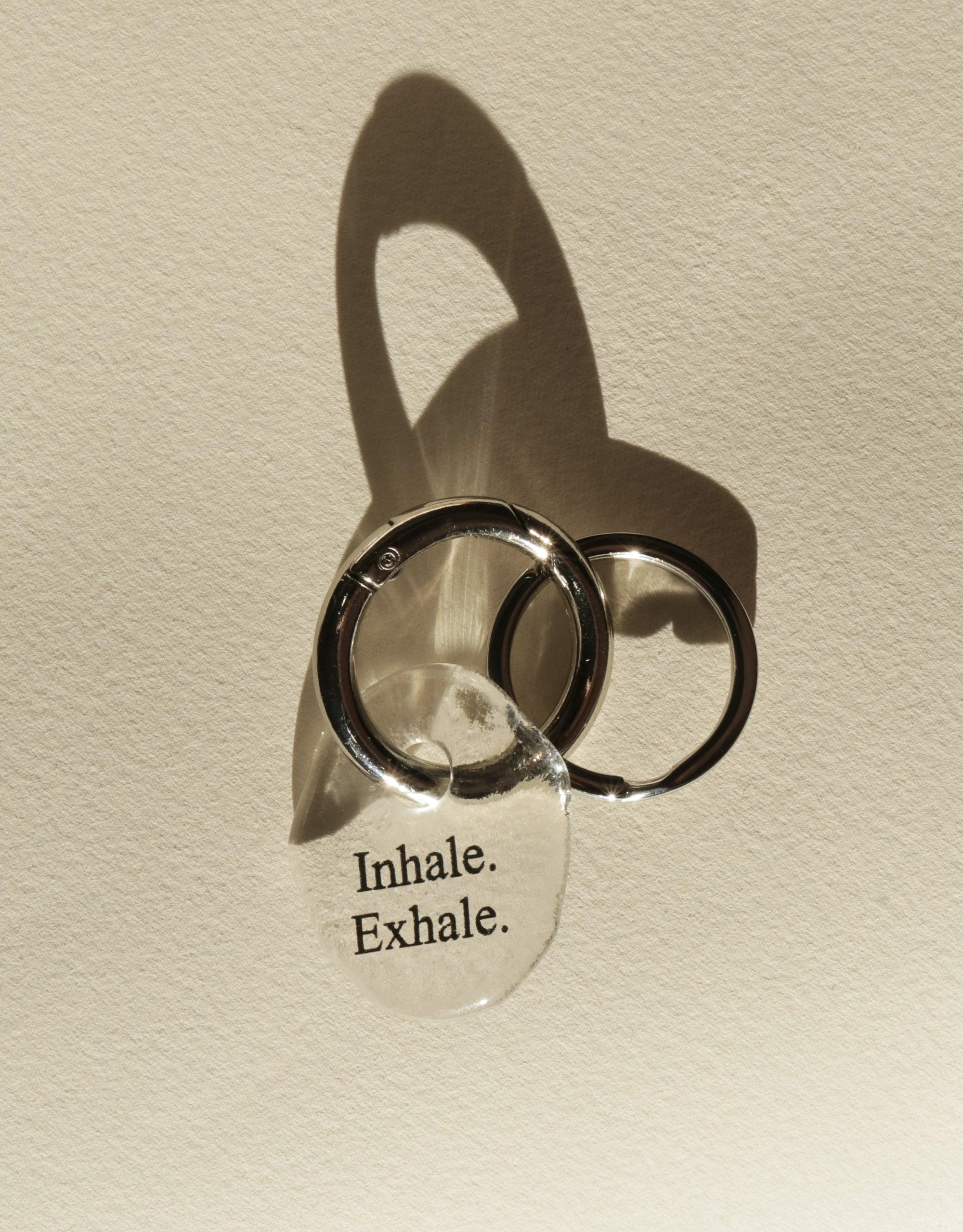 Messenger Keyring with "Inhale. Exhale." engraved on clear glass charm, attached to stainless steel rings. Eco-friendly repurposed glass keychain casting shadow on beige background. Stylish and mindful accessory from the Messenger Collection by CLED.