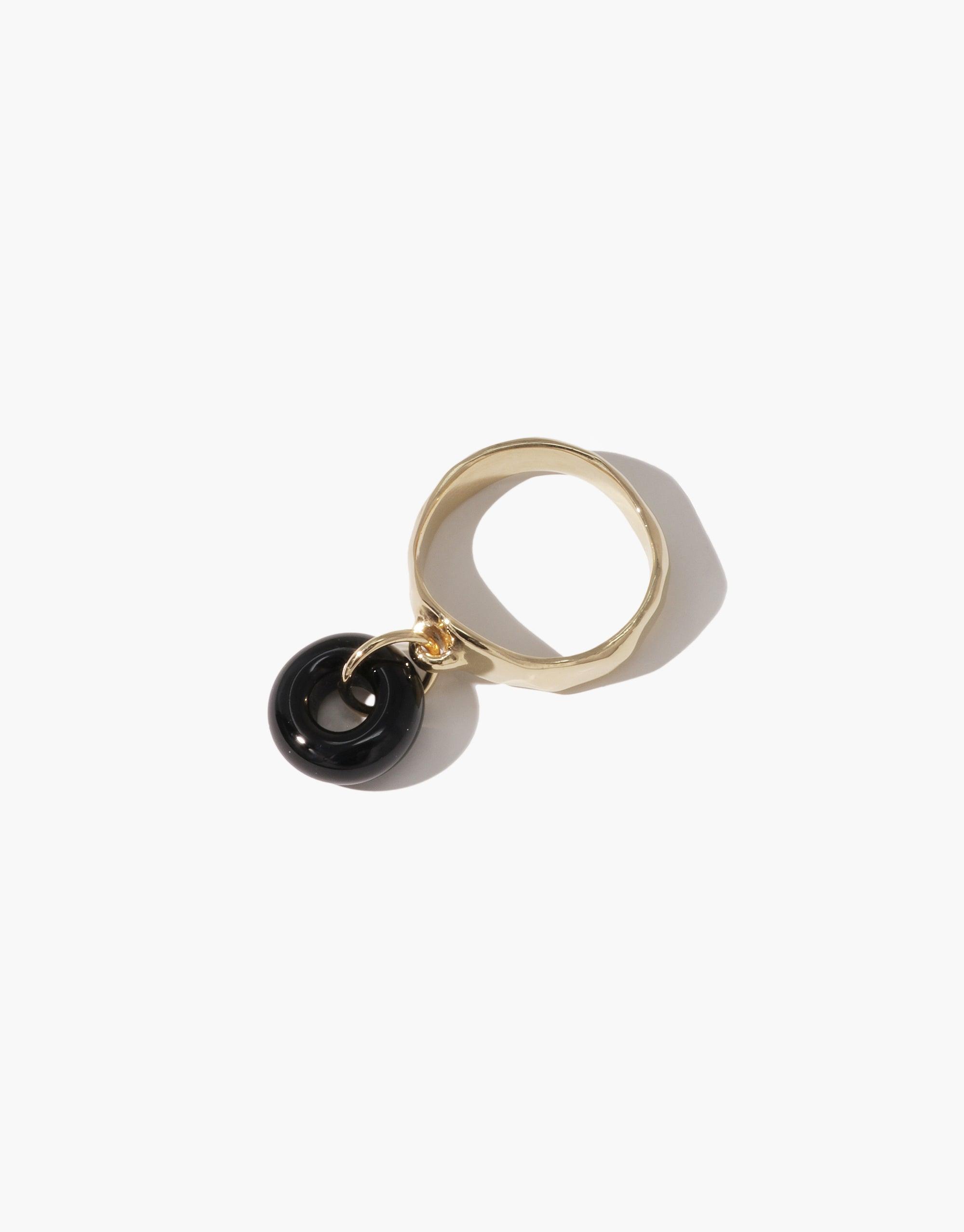 Gold-toned ring with dangling black circular charm, showcasing the Donut Dangling Ring. Elegant and playful design, perfect for eco-conscious jewelry lovers. Handcrafted with upcycled materials. CLED