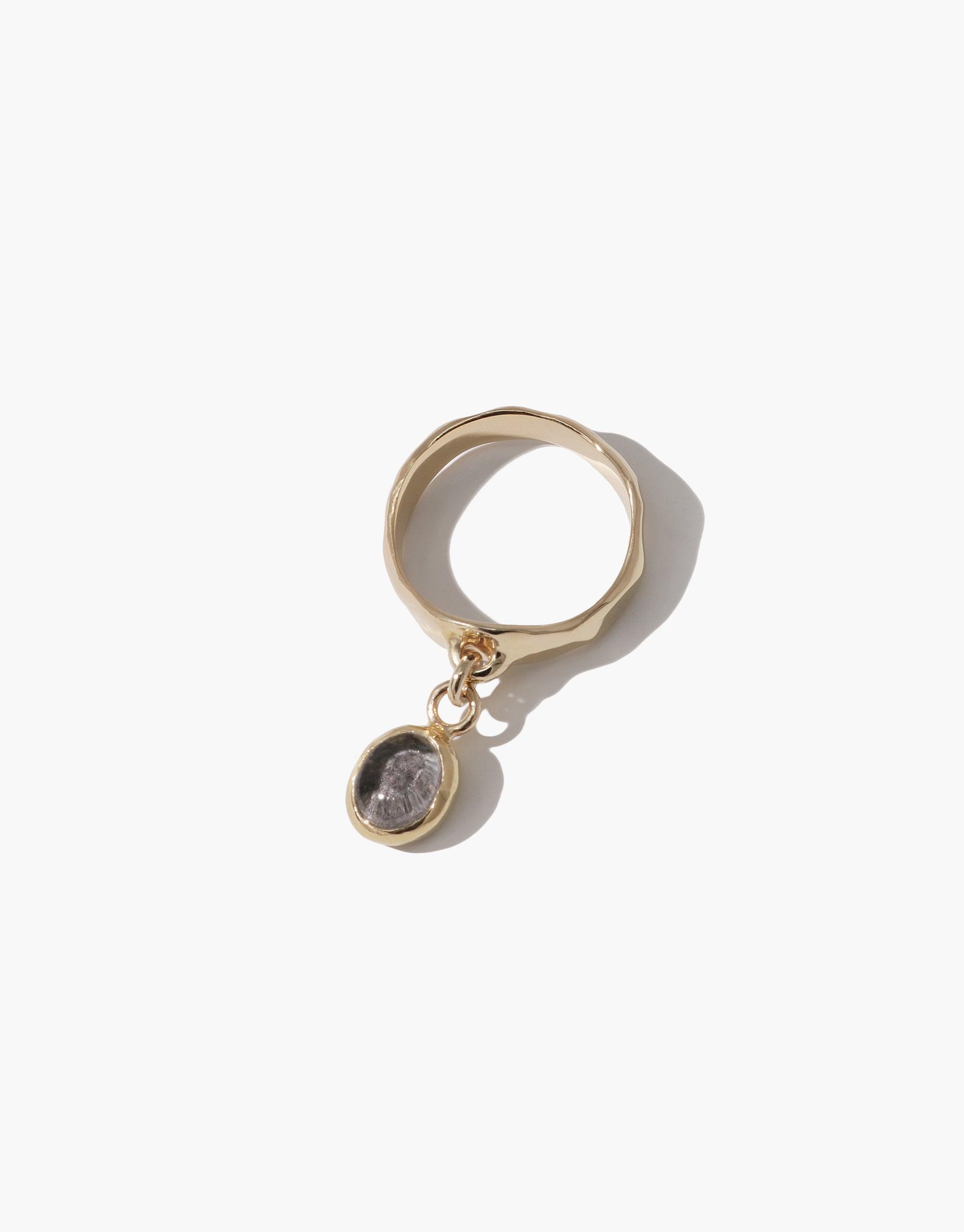 Oval Dangling Ring featuring a gold-toned band with a suspended circular charm. The charm contains a gray upcycled eco gem, showcasing sustainable design. Handcrafted jewelry piece with minimalist elegance. CLED