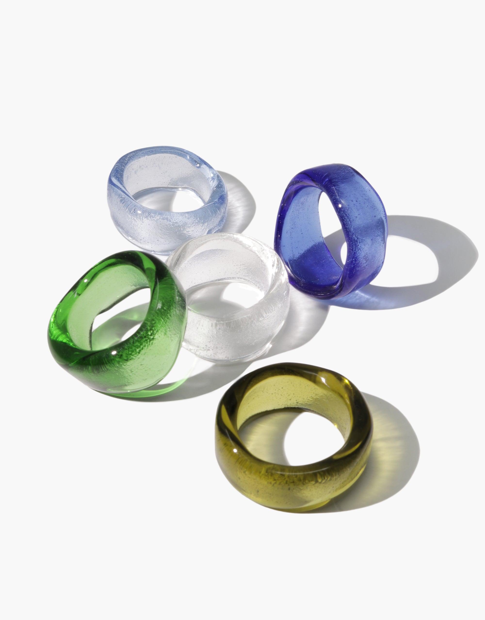 Wave Ring: A collection of eco-conscious rings made from discarded glass bottles, showcasing hand-formed designs in light olive, clear, green, light blue, and blue, arranged on a white surface.