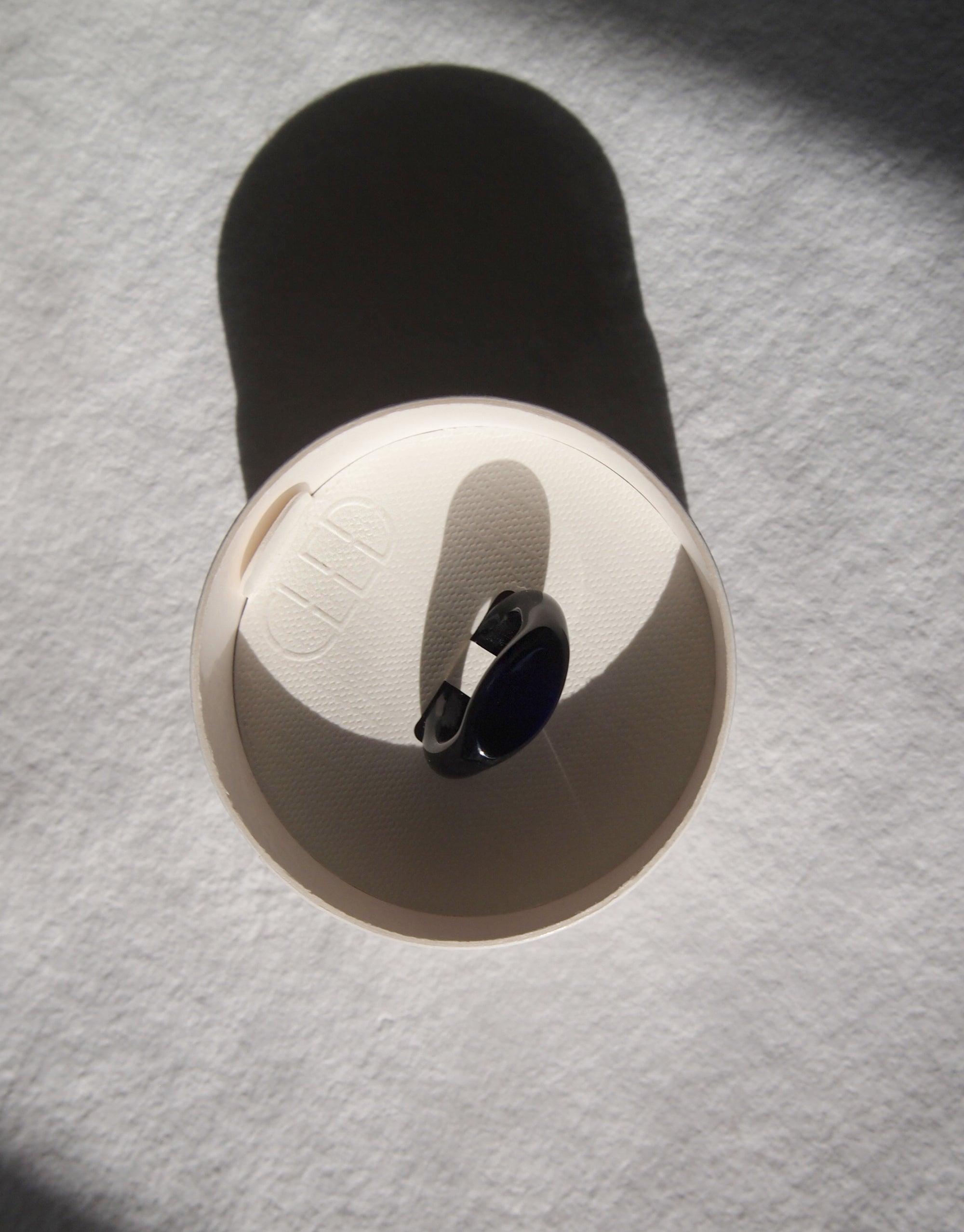 Signet Ring Original by CLED, showcasing a sleek black eco gem set in a circular metal band, casting a dramatic shadow on a white surface. Elegantly crafted from repurposed glass, this sustainable jewelry piece embodies eco-conscious design.