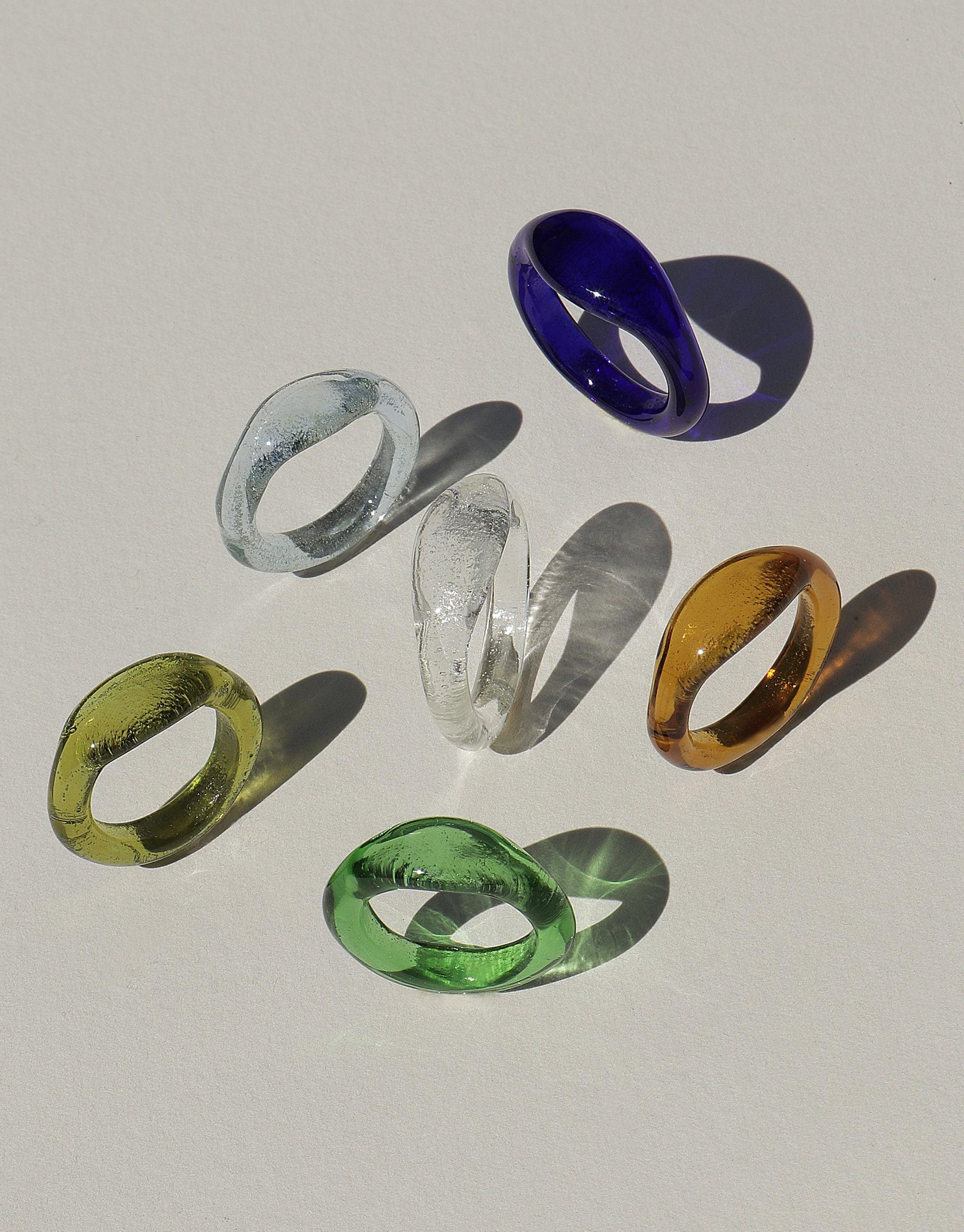 Six colorful eco-friendly signet rings made from repurposed glass, displayed on a light background. Rings showcase various hues including blue, green, yellow, and clear. Part of the Signet Ring Original collection by CLED.