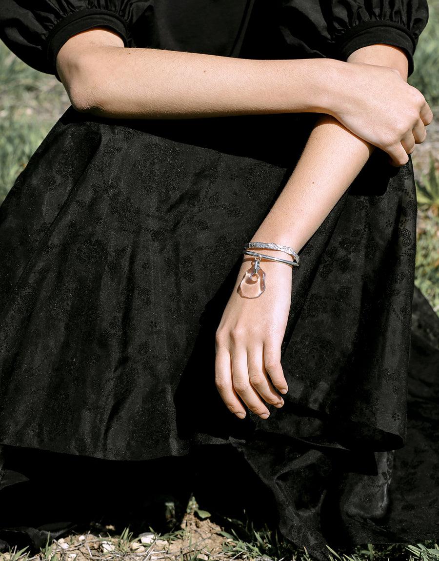 Elegant silver Cliff Bracelet on wrist, showcasing simple curved design against black fabric background. Handcrafted sterling silver cuff bracelet with earthy aesthetic. Versatile accessory for any outfit. CLED