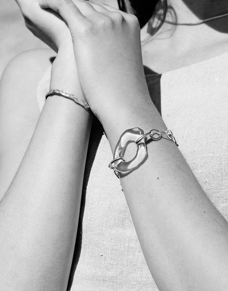 Black and white image of hands wearing elegant bracelets, including a simple curved cuff bracelet. Showcasing the Cliff Bracelet's minimalist design and versatile style. Handcrafted sterling silver jewelry by CLED.