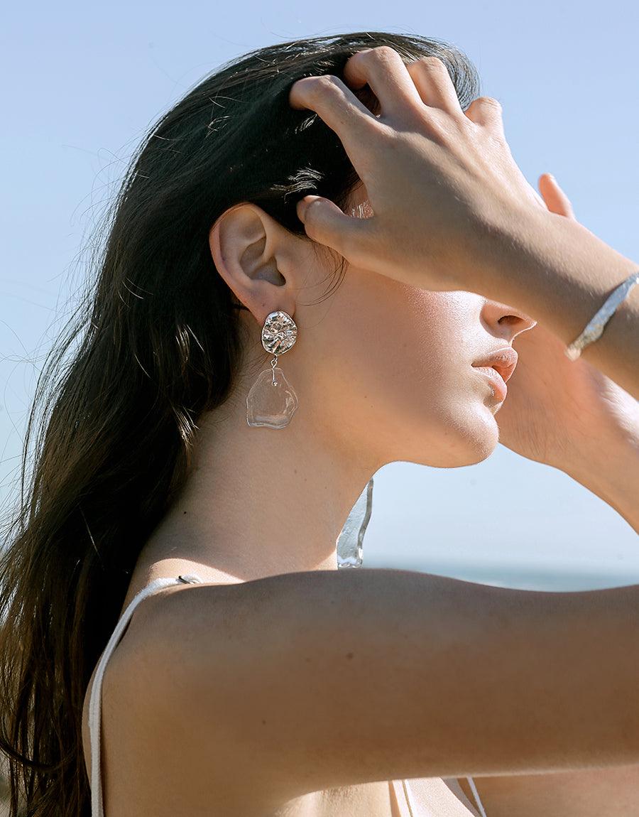 Elegant model showcasing Coastline Earrings | Small Drop, featuring a clear eco-glass pendant and silver stud against a blue sky backdrop. Hand-crafted sustainable jewelry inspired by coastal shapes, perfect for a stylish and eco-conscious look. CLED.