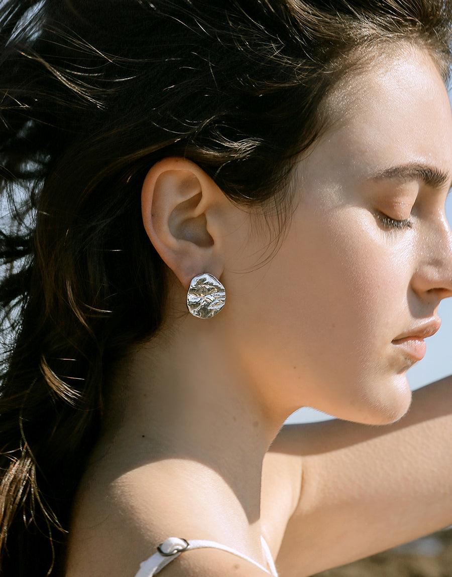 Elegant model wearing Coastline Earrings | Small Drop, showcasing textured silver stud with eco-friendly glass design. Earring reflects light, complementing model's profile and dark hair. Handcrafted sustainable jewelry by CLED.