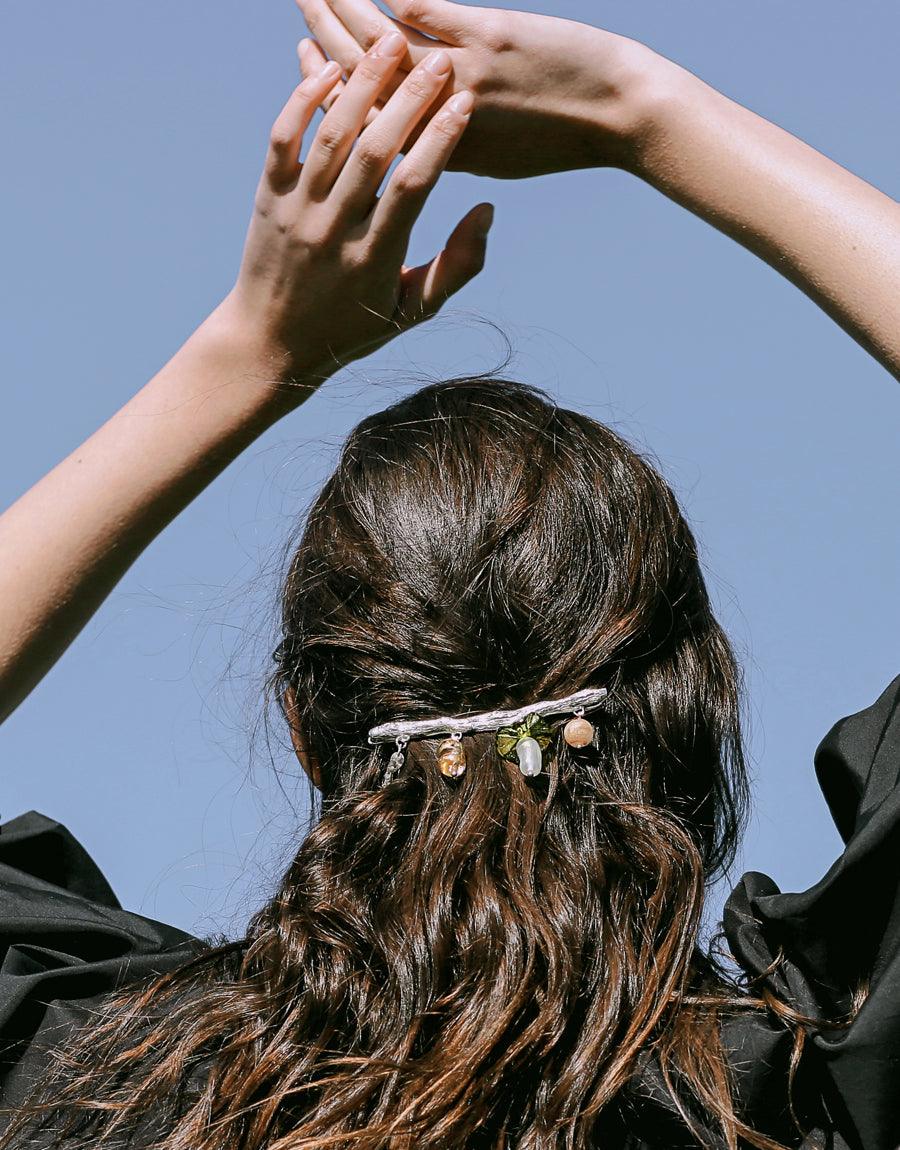 Elegant Floral Barrette adorning long, wavy dark hair against blue sky. Model's raised arms frame the hair accessory, showcasing its delicate design and eco-friendly charm. Perfect for spring and summer styling. Handcrafted sterling silver with repurposed glass elements. CLED Floral Barrette.