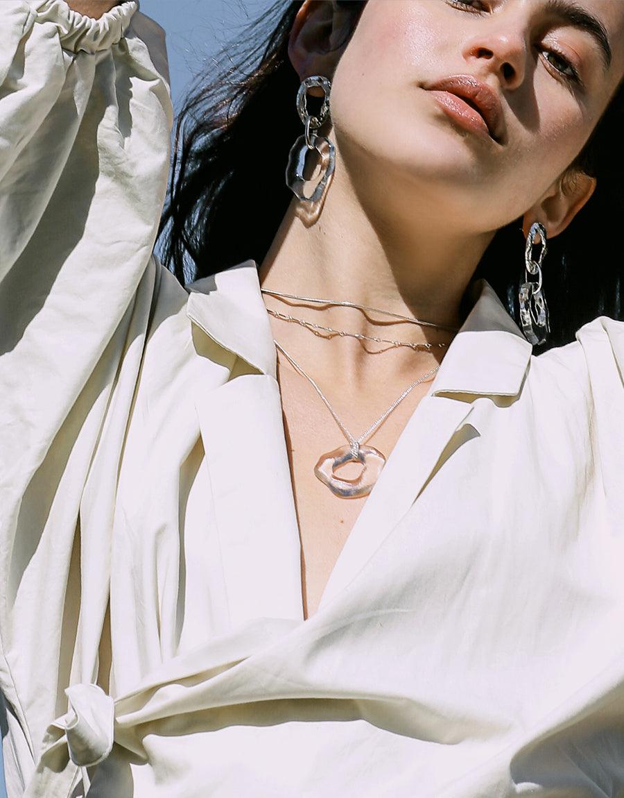 Elegant model wearing High Tide Earrings, featuring sculptural silver and eco-glass design. Paired with white blouse and delicate necklace, showcasing versatile ocean-inspired statement piece. Sunlit portrait highlights earrings' unique handcrafted aesthetic. CLED High Tide Earrings.