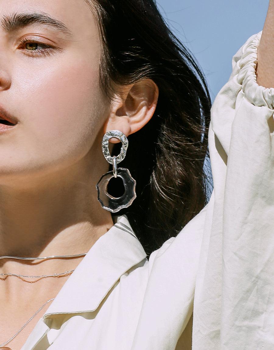 Elegant High Tide Earrings featuring a double-drop design with circular and wave-like shapes, crafted from sterling silver and eco-friendly repurposed glass. Displayed on a model wearing a white outfit against a blue background, showcasing the earrings' statement style. CLED