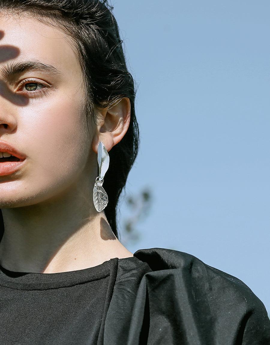 Close-up of elegant Leaf Earrings, showcasing delicate sterling silver design with eco-friendly repurposed glass pendant. Sunlight illuminates the intricate botanical-inspired shape, highlighting its shimmer against a blue sky background. Handcrafted sustainable jewelry by CLED.