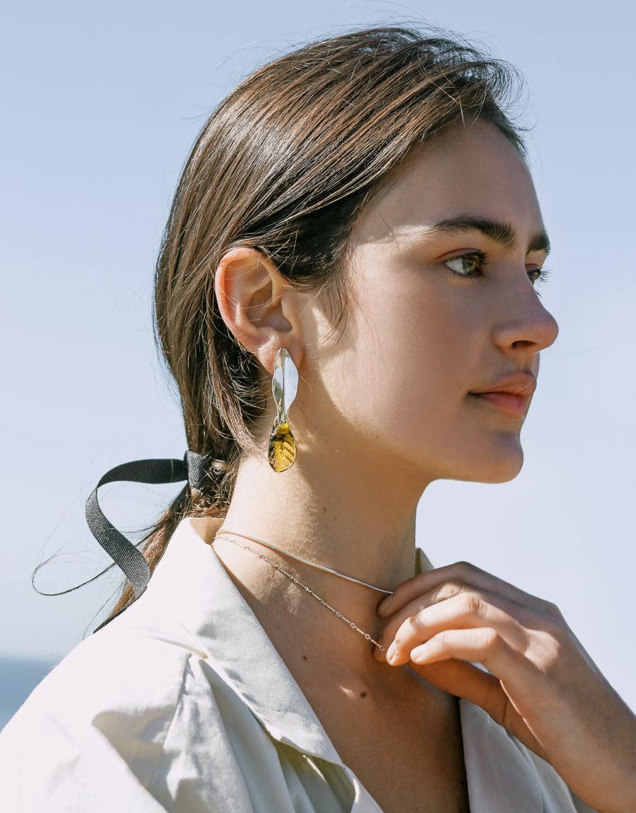 Close-up side profile of a model wearing elegant Leaf Earrings, featuring a delicate silver stem and a translucent amber-colored leaf-shaped pendant, showcasing the earring's botanical inspiration and handcrafted design. Leaf Earrings by CLED.