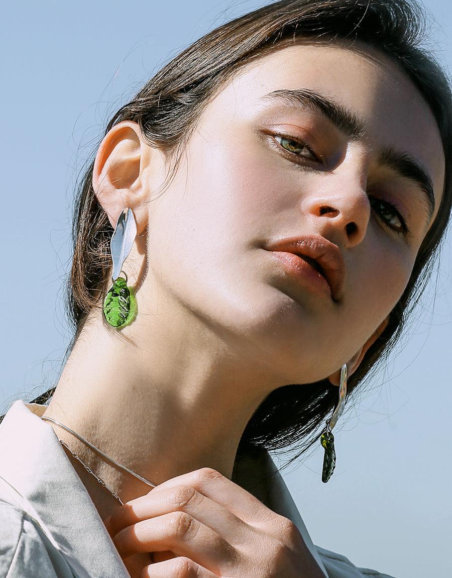 Elegant Leaf Earrings showcasing sustainable design with hand-formed eco gems in vibrant green, dangling from sterling silver hooks. Model wearing earrings against blue sky background, highlighting the botanical-inspired jewelry's shimmering effect. Handcrafted by CLED.