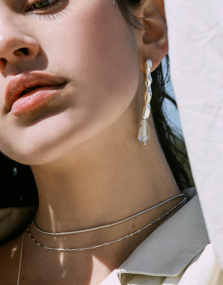 Water Earrings: Artistic asymmetric earrings featuring wavy, free-formed shapes, handcrafted from repurposed glass bottles, highlighting eco-conscious design. Sold in sterling silver, measuring 2 to 2.5 inches in length.