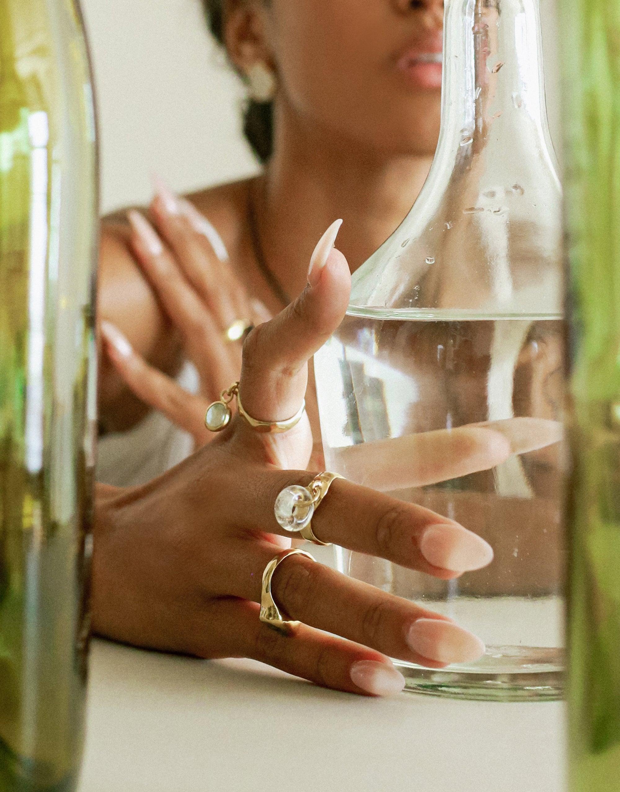 Hand adorned with delicate gold rings and Donut Dangling Ring, grasping clear glass bottle. Soft focus on elegant jewelry pieces showcasing eco-friendly design. Artistic composition highlights sustainable luxury accessories. CLED Donut Dangling Ring.